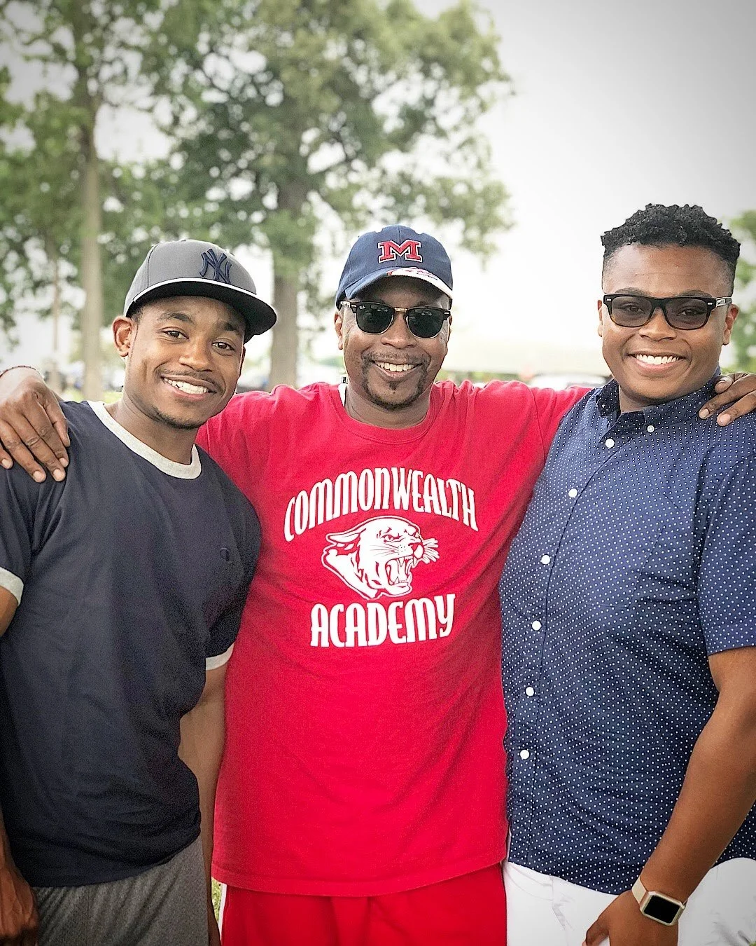 Three men smiling with their arms around each other outdoors, wearing casual clothing and sunglasses. The man in the middle wears a red Commonwealth Academy t-shirt and a baseball cap, the man on the left wears a black NY Yankees cap, and the man on 