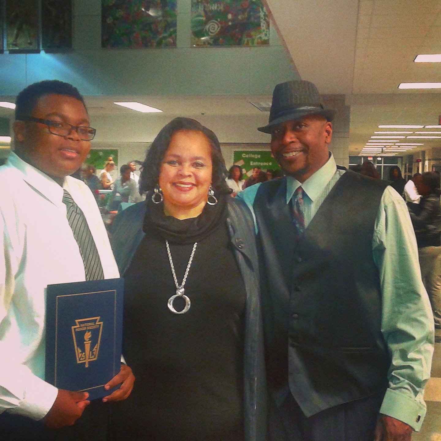 Three people standing together at an indoor event, two men and one woman, smiling and dressed in formal attire.