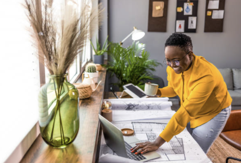 Person smiling while working on architectural plans at a desk, with a laptop and coffee mug in hand. A green vase with decorative grass and a potted plant are on the windowsill.