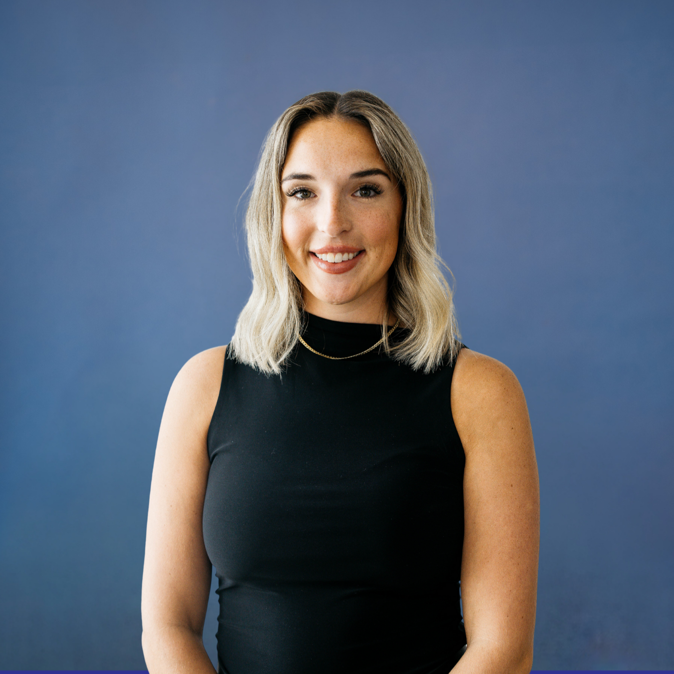 A woman with shoulder-length blonde hair wearing a black sleeveless top and a gold chain necklace, smiling against a plain blue background.