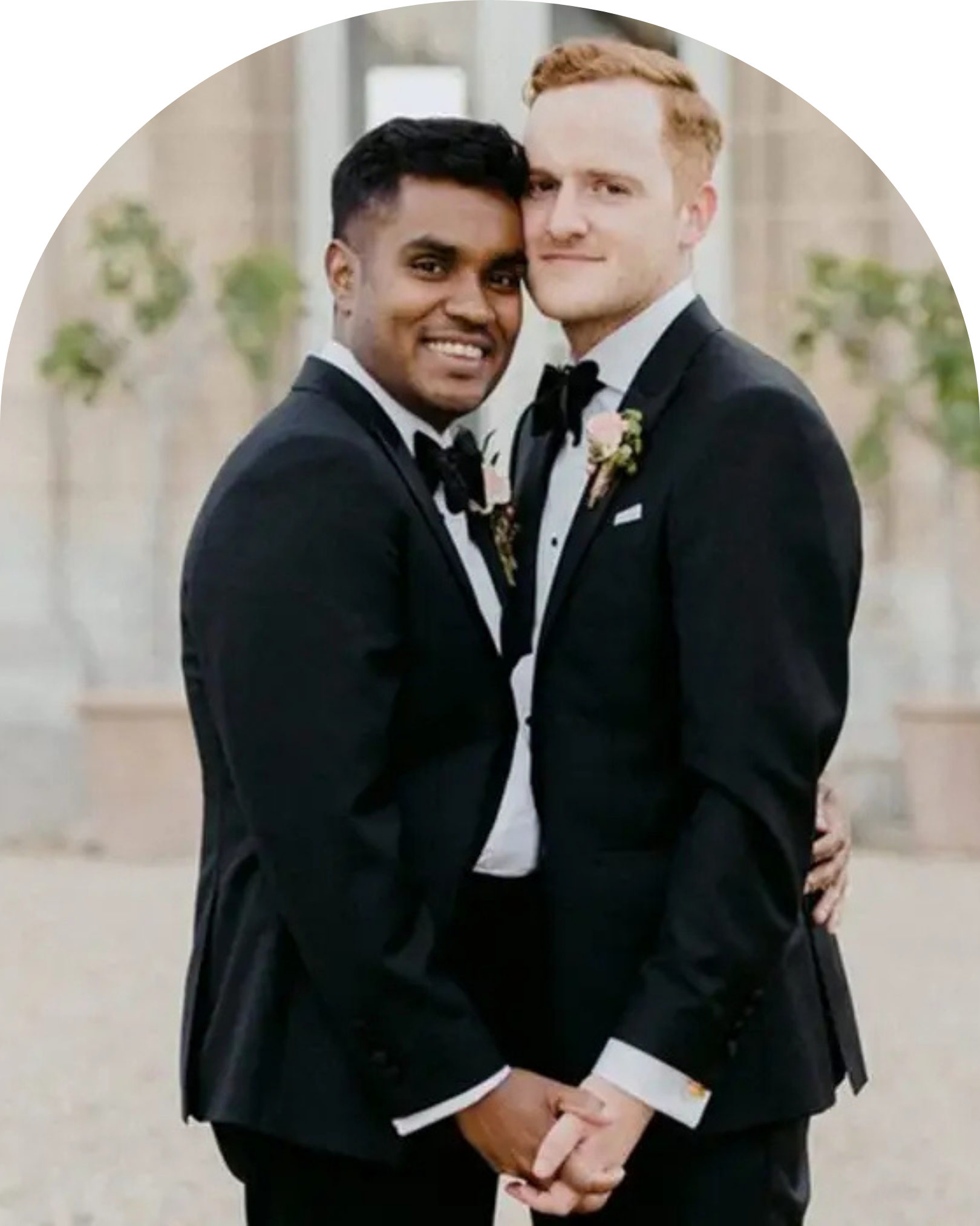 Two men in tuxedos, embracing and smiling for a photo at a wedding, with a blurred architectural background and greenery.