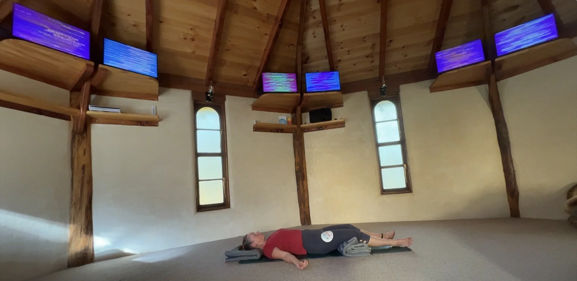 Tara lying on back on yoga mat in meditation room with wooden beams and windows, overhead are six EESystem units.