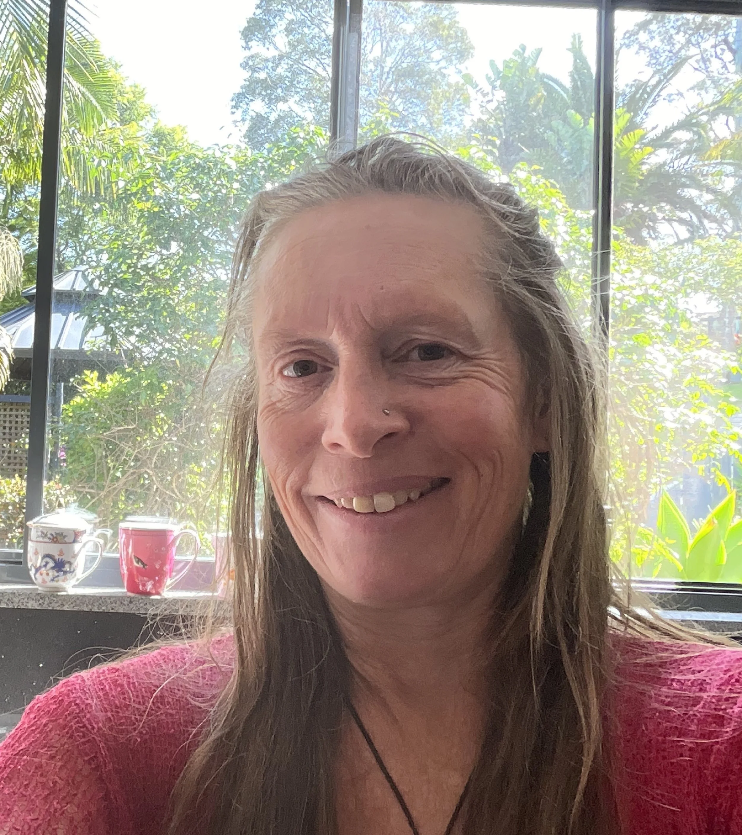 Tara with long hair smiling, sitting inside near a window with plants and two colorful cups on the windowsill, sunny outdoor scene in the background.