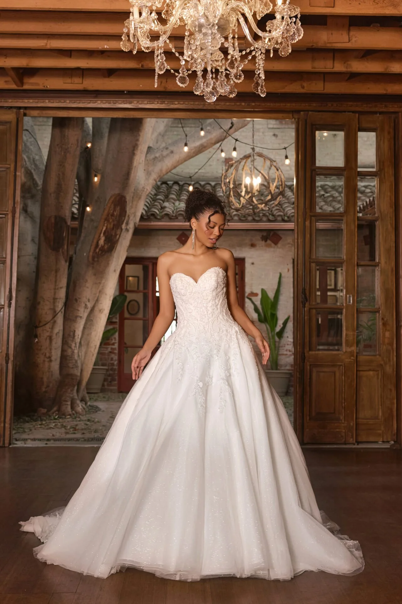A woman wearing a white wedding gown with lace details, standing in a rustic indoor setting with wooden elements, plants, and chandeliers.