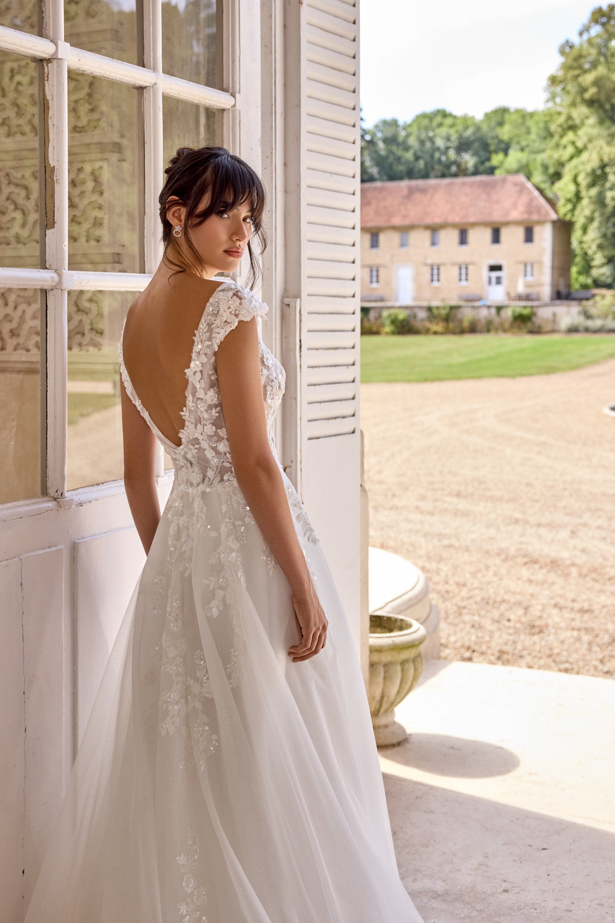 A woman in a white lace wedding dress standing by an open window outside a building with a red-tiled roof, trees, and a green lawn in the background.