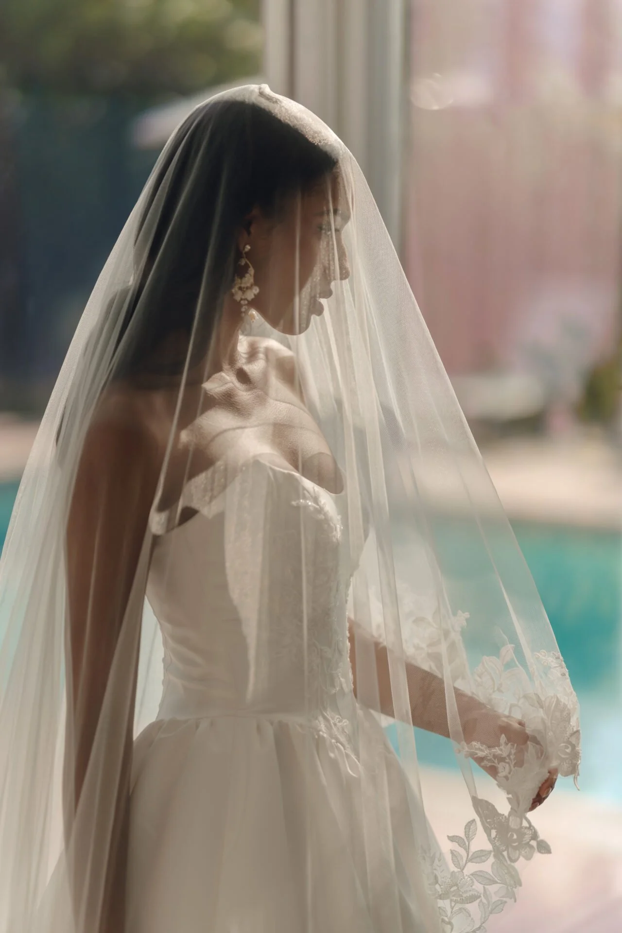 A woman wearing a Stella York wedding dress and veil standing near a window, with sunlight illuminating her profile, holding a bouquet of flowers.