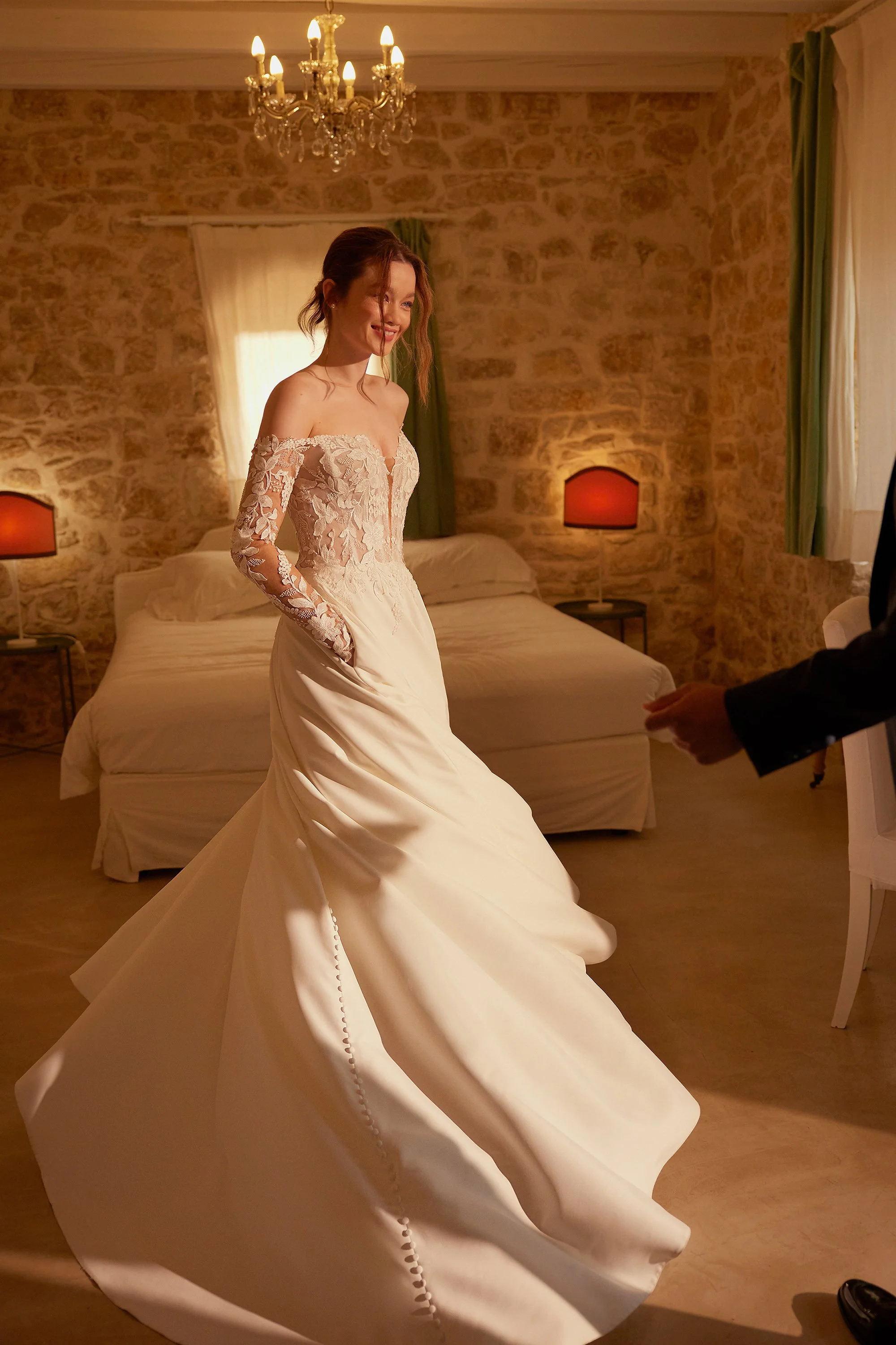 A bride in a white lace off-the-shoulder wedding dress standing in a warmly lit bedroom with stone walls, a chandelier, and bedside lamps.