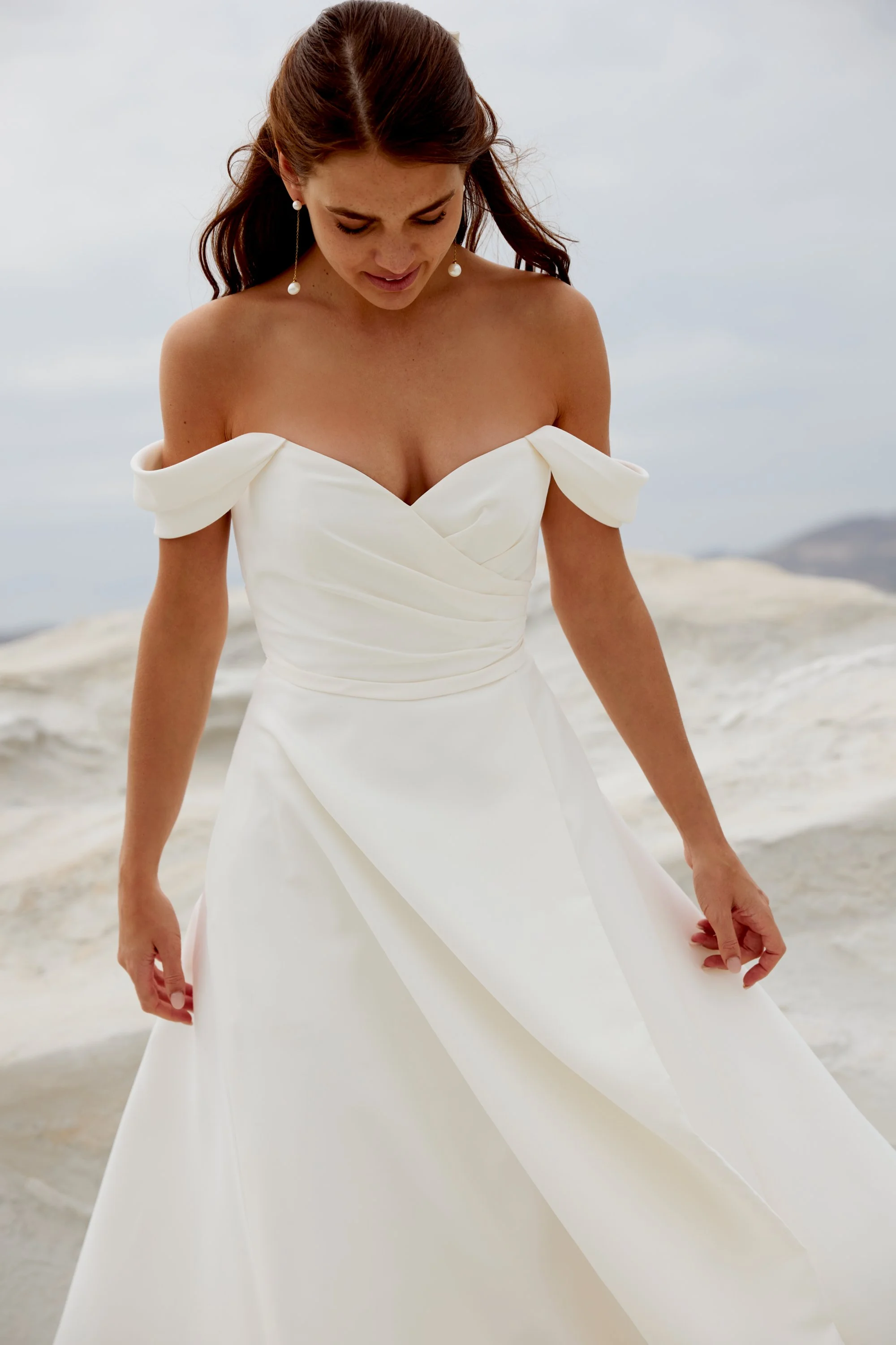 A woman in a white off-shoulder gown with bow details on the sleeves, standing on a beach. She has long brown hair and is wearing long pearl earrings. The background shows a cloudy sky and distant mountains.