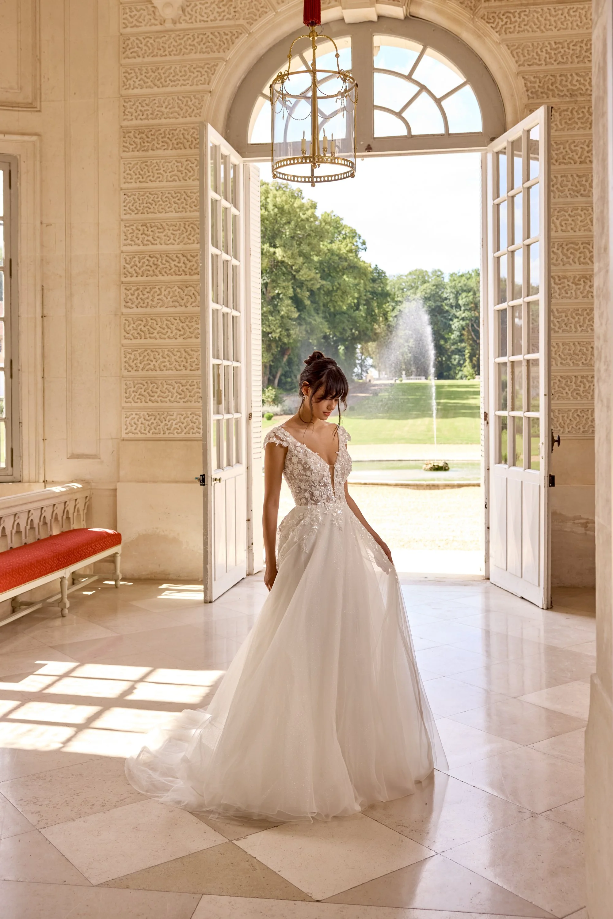 A bride in a white wedding gown standing in a bright, elegant room with open French doors leading outside to a lush green lawn and fountain, sunlight casting shadows on the tiled floor.