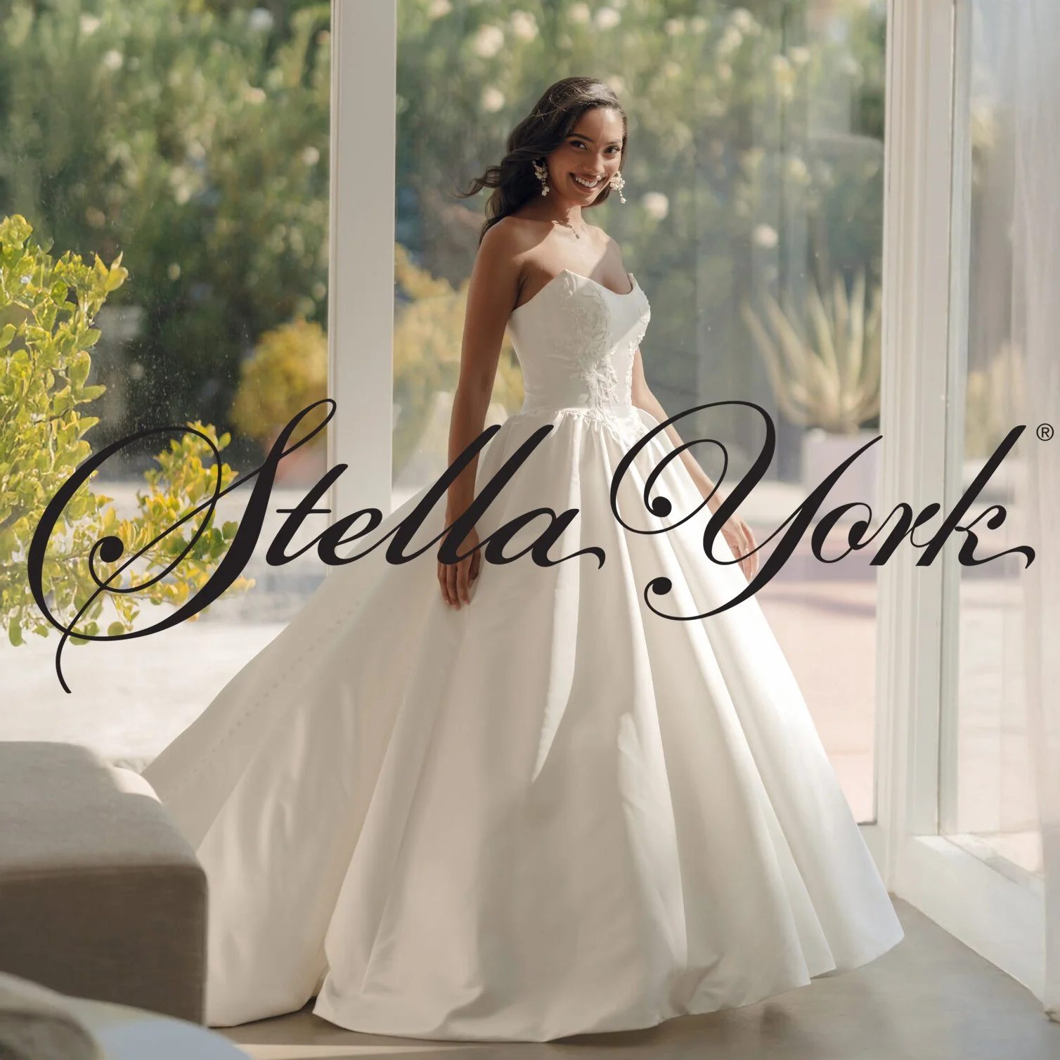 A woman in a white wedding gown standing by a large window with sunlight. The dress has a fitted bodice and a full skirt. She is smiling and looking at the camera. The logo 'Stella York' is overlaid on the image.