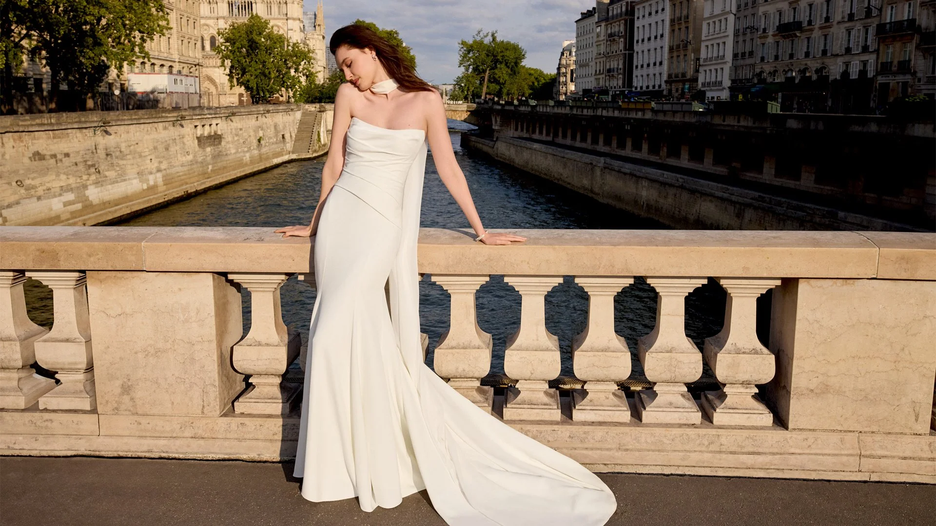 Woman in a strapless white dress with a train, standing on a stone bridge railing over a river in an urban area with historic buildings in the background.