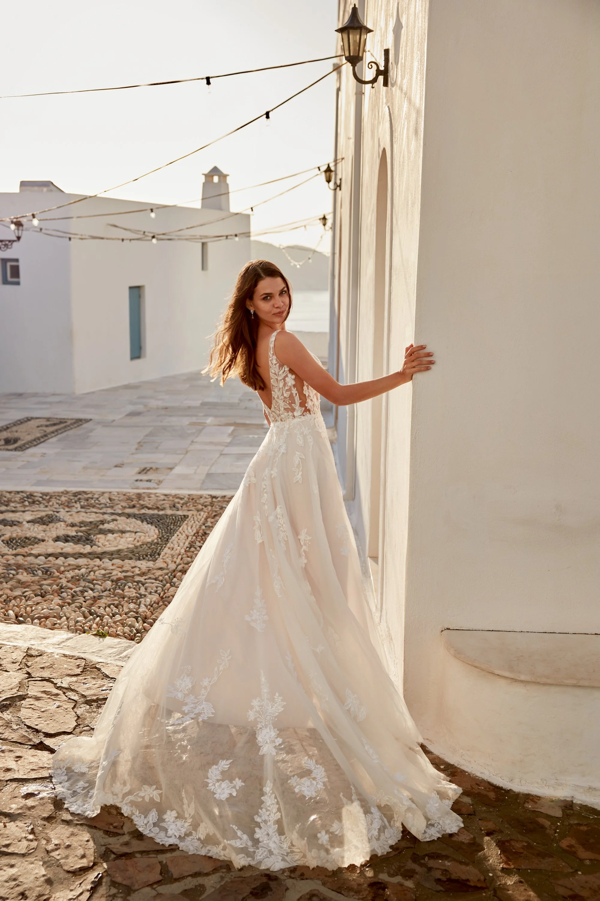 A woman in a white wedding dress stands outdoors near a white wall with her hand touching it, and looks back over her shoulder at the camera in the late afternoon or early evening.