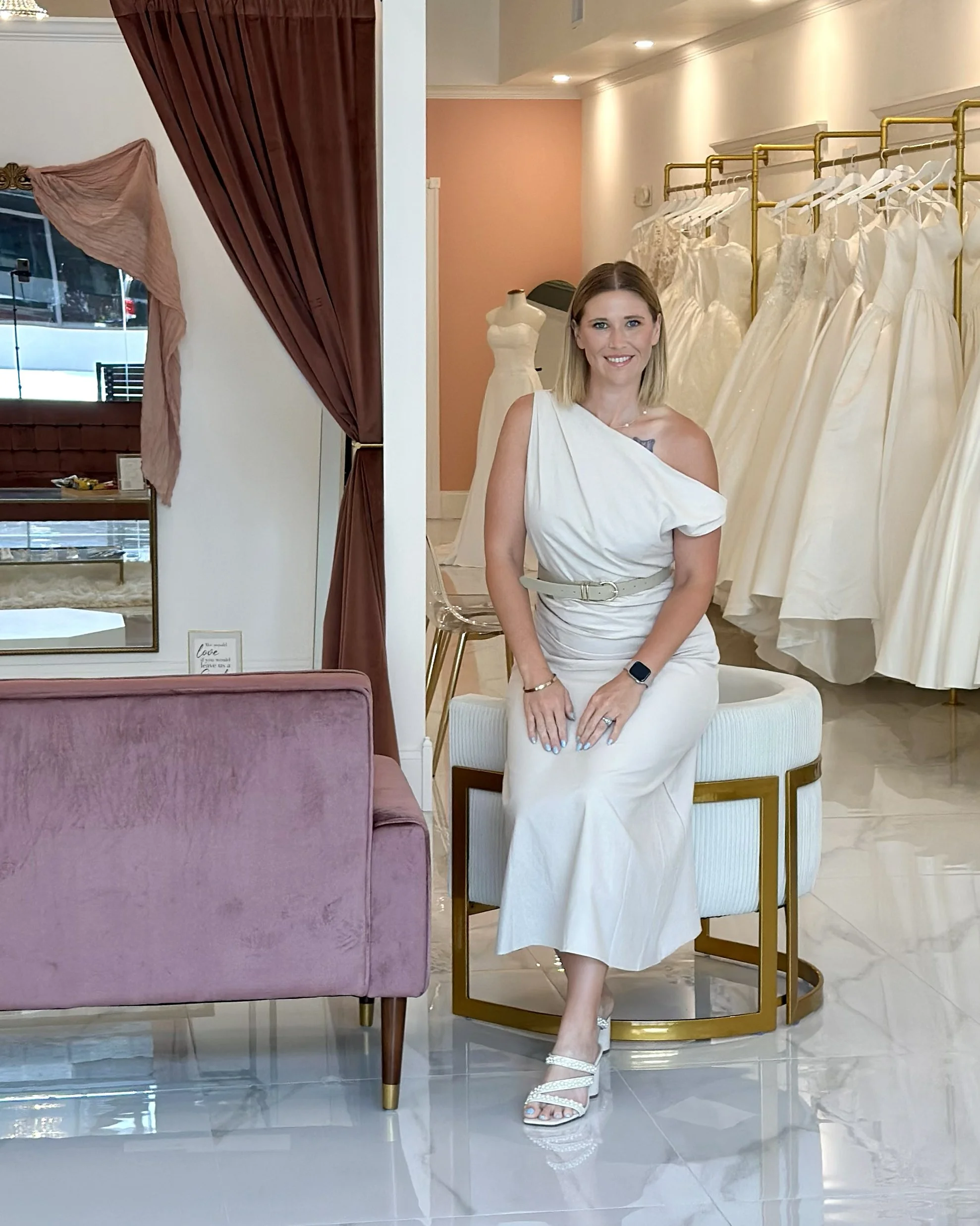 A woman in a white dress sitting on a gold and white stool in The Luxe Bride Boutique, surrounded by wedding dresses on racks, with a pink wall and a mannequin in the background.