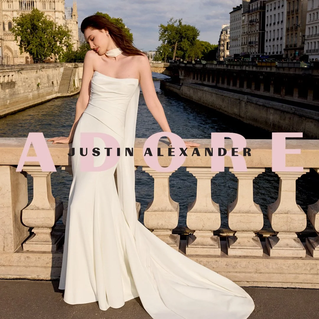 A woman in a white strapless gown standing on a bridge over a river in a city, with the title 'ADORE' and the name 'Justin Alexander' overlaid.