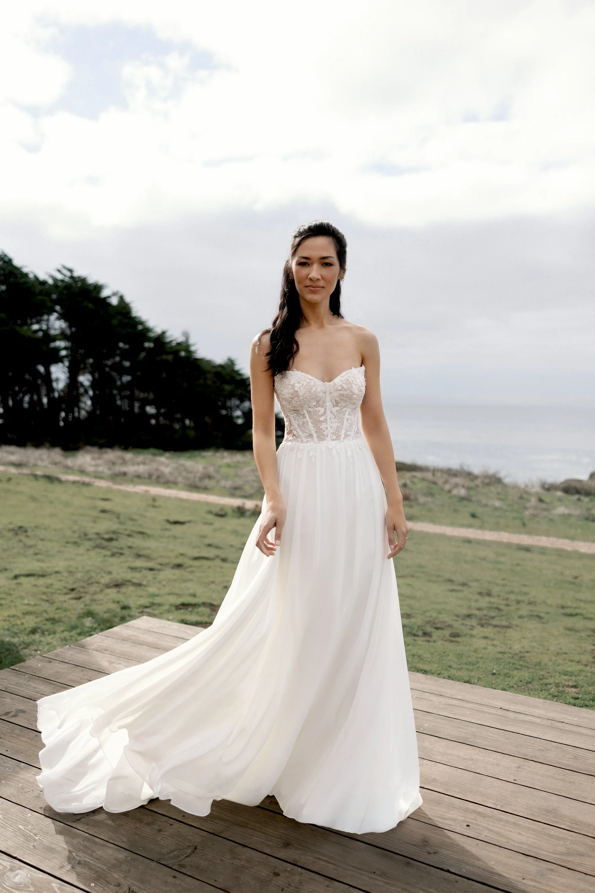A woman in a strapless white wedding dress with lace bodice standing on a wooden platform outdoors with grassy fields and trees in the background.