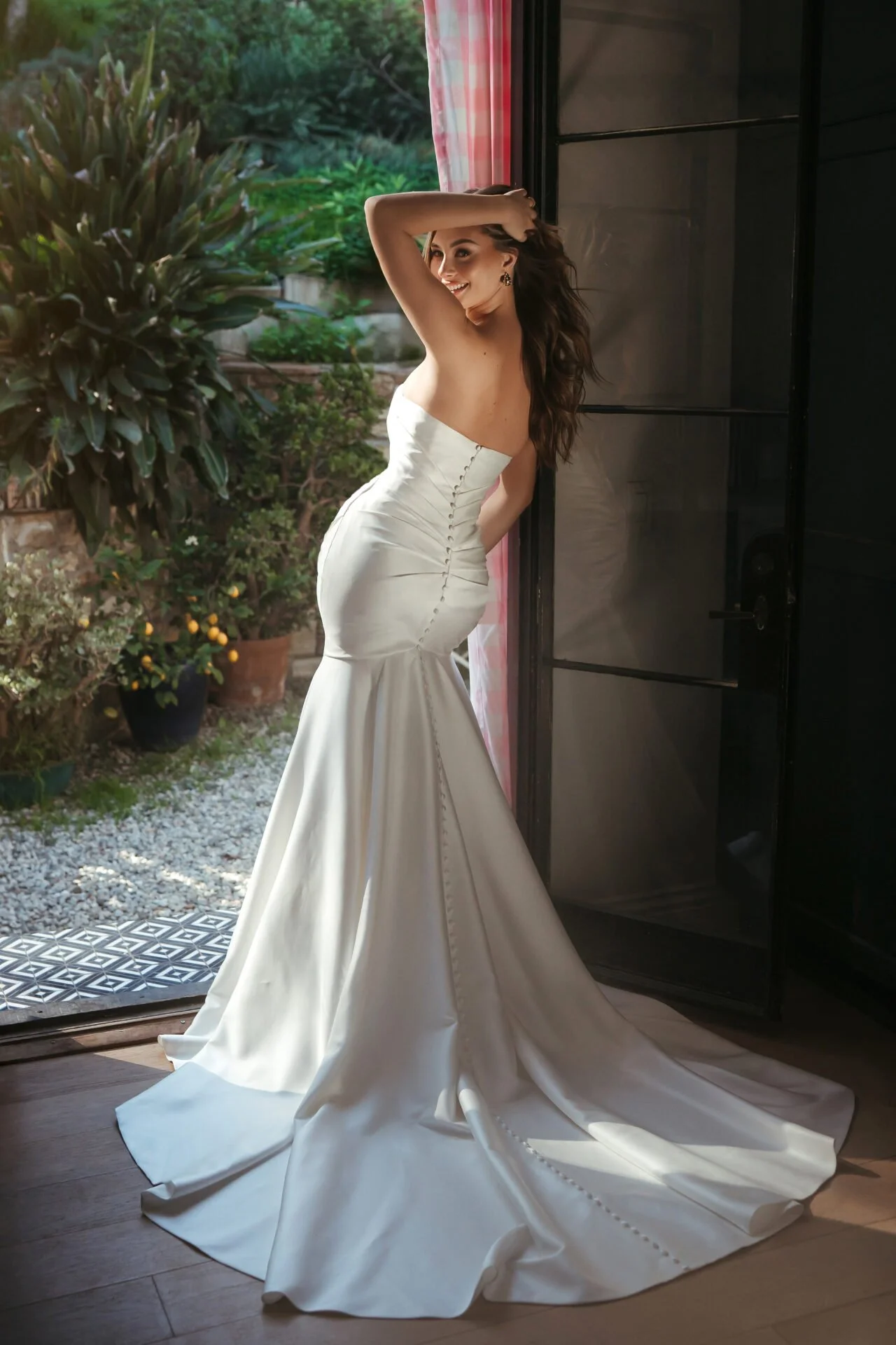 A woman in a white Stella York strapless wedding dress with a long train stands near an open door with colorful curtains, smiling and touching her hair. Outside, there are potted plants and greenery.