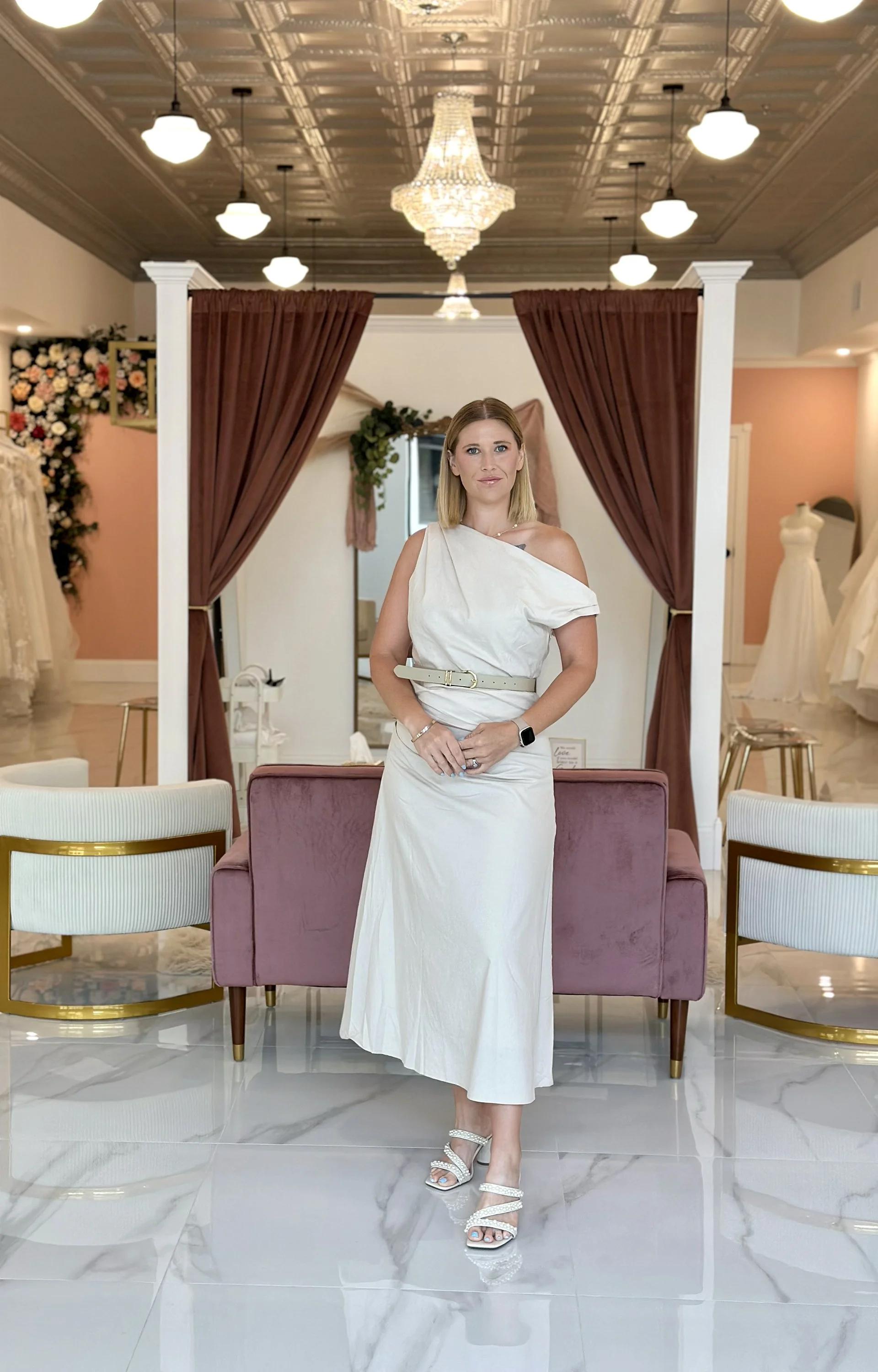 A woman wearing a white dress, silver jewelry, and high-heeled sandals standing in a luxurious Luxe Bride Boutique showroom with elegant decor, chandeliers, curtains, and wedding dresses in the background- serving Central Florida brides.