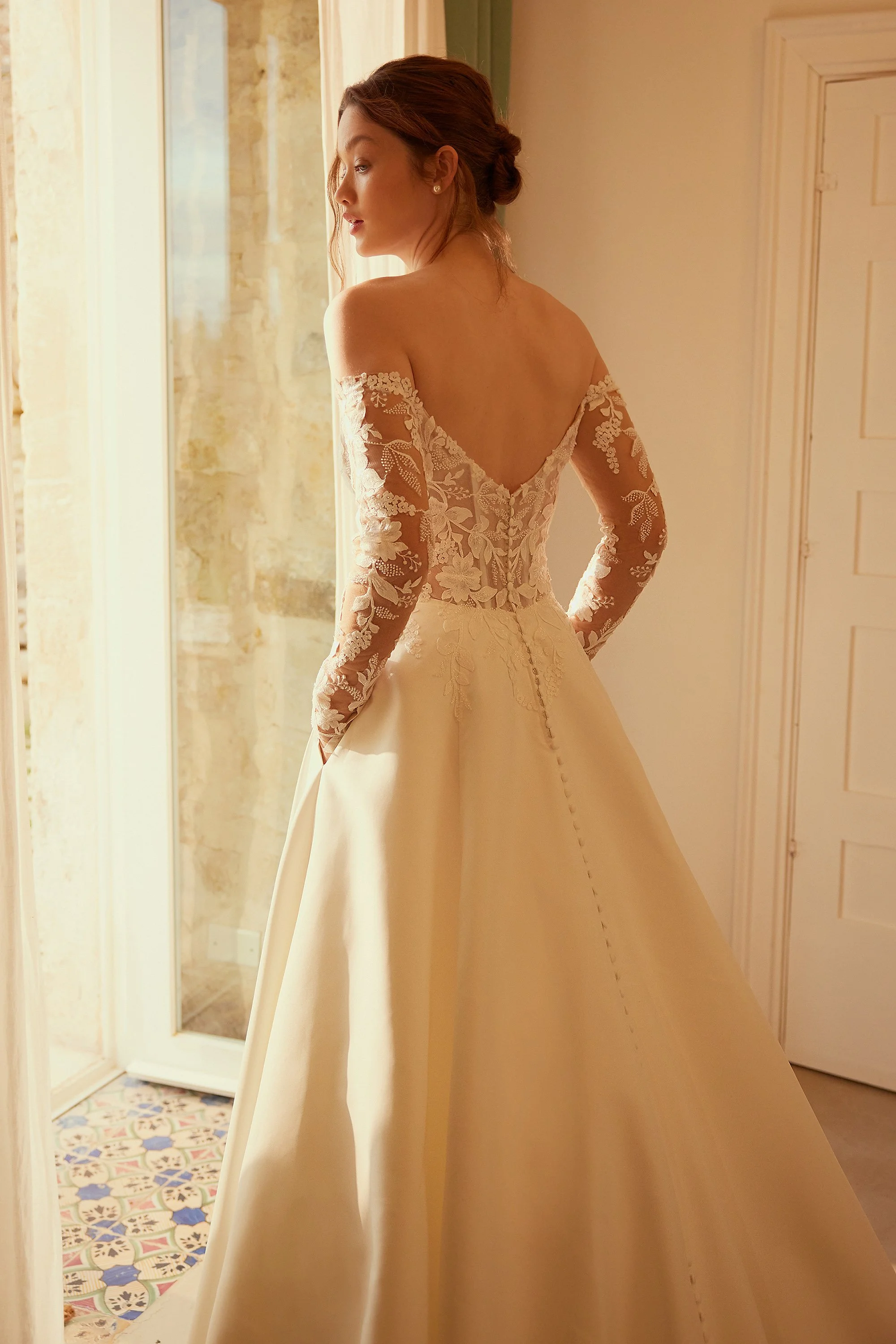 A woman in a wedding dress standing indoors near a window, looking to her left, with a patterned tile floor visible outside a glass door.