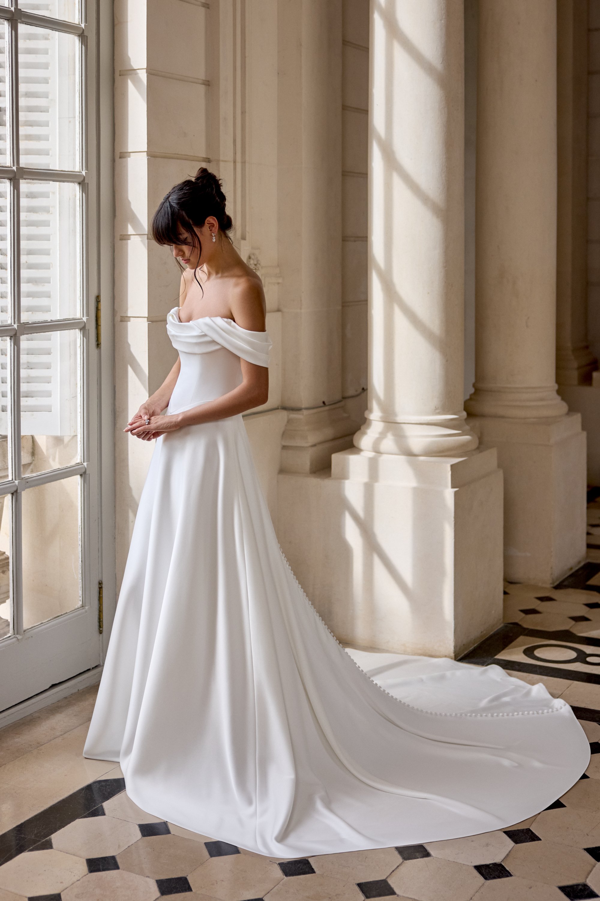 A woman in a white wedding dress standing near a large window with sunlight streaming in, with classical columns in the background.