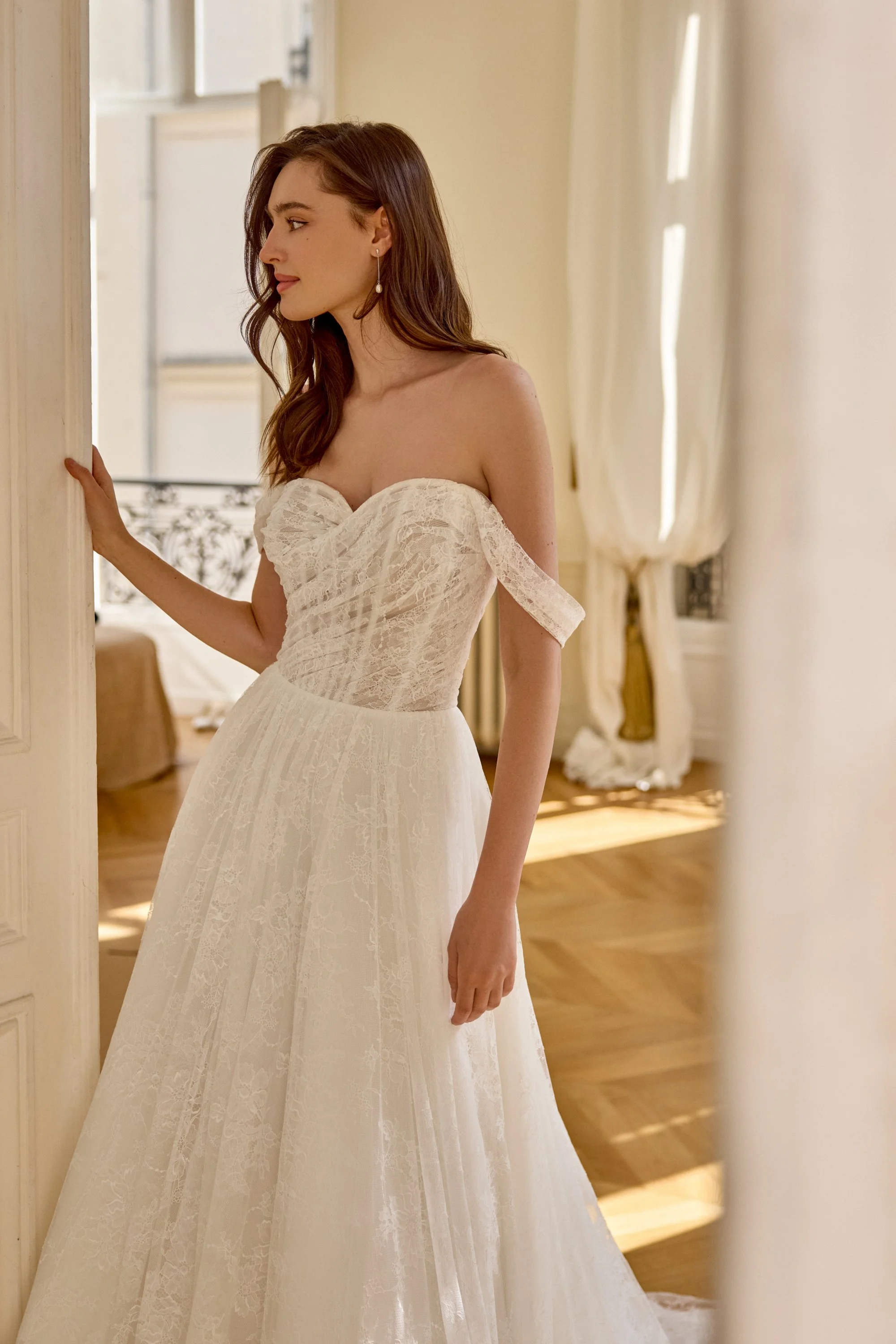 A woman in a wedding dress standing inside a room, holding onto the door frame and looking to the side.