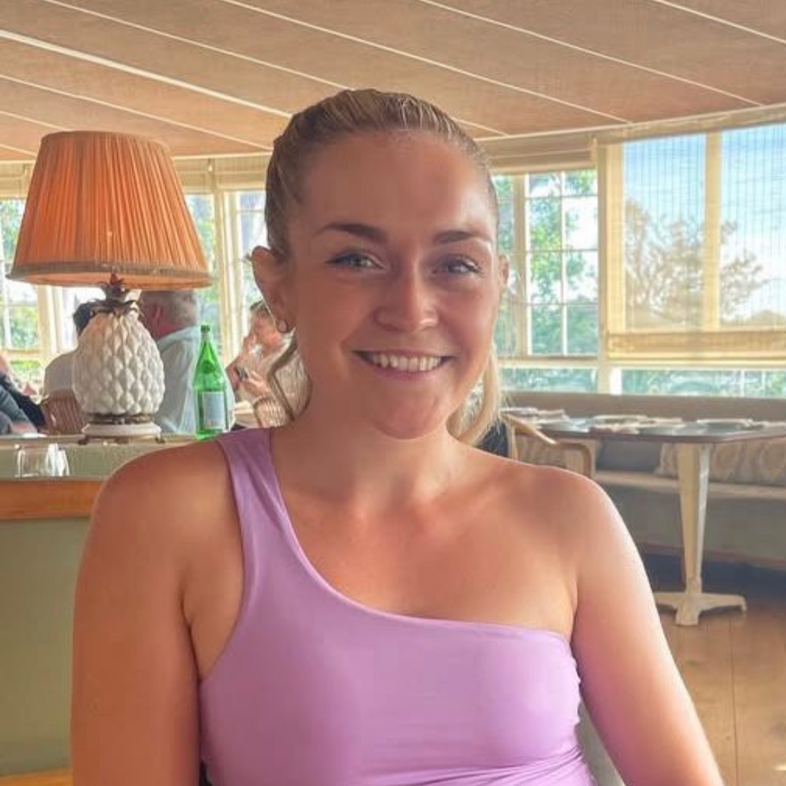 A smiling woman with blonde hair tied back, wearing a purple sleeveless top, sitting indoors near large windows with a view of greenery outside.