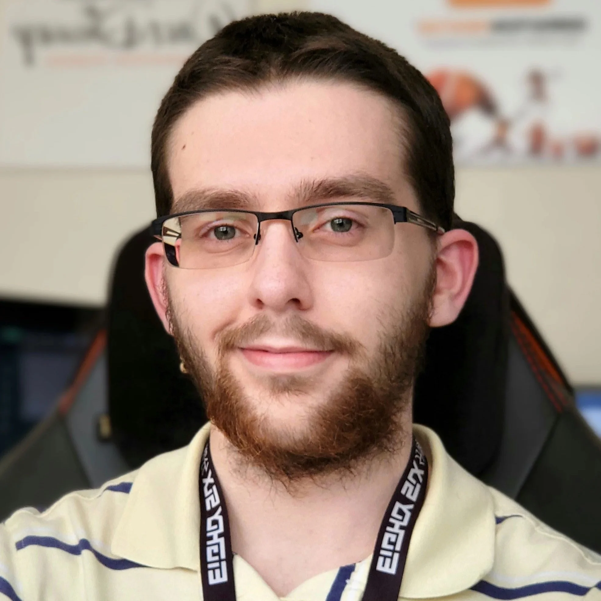 A young man with glasses, a beard, and short dark hair taking a selfie, sitting in a chair with a gaming or office environment in the background.