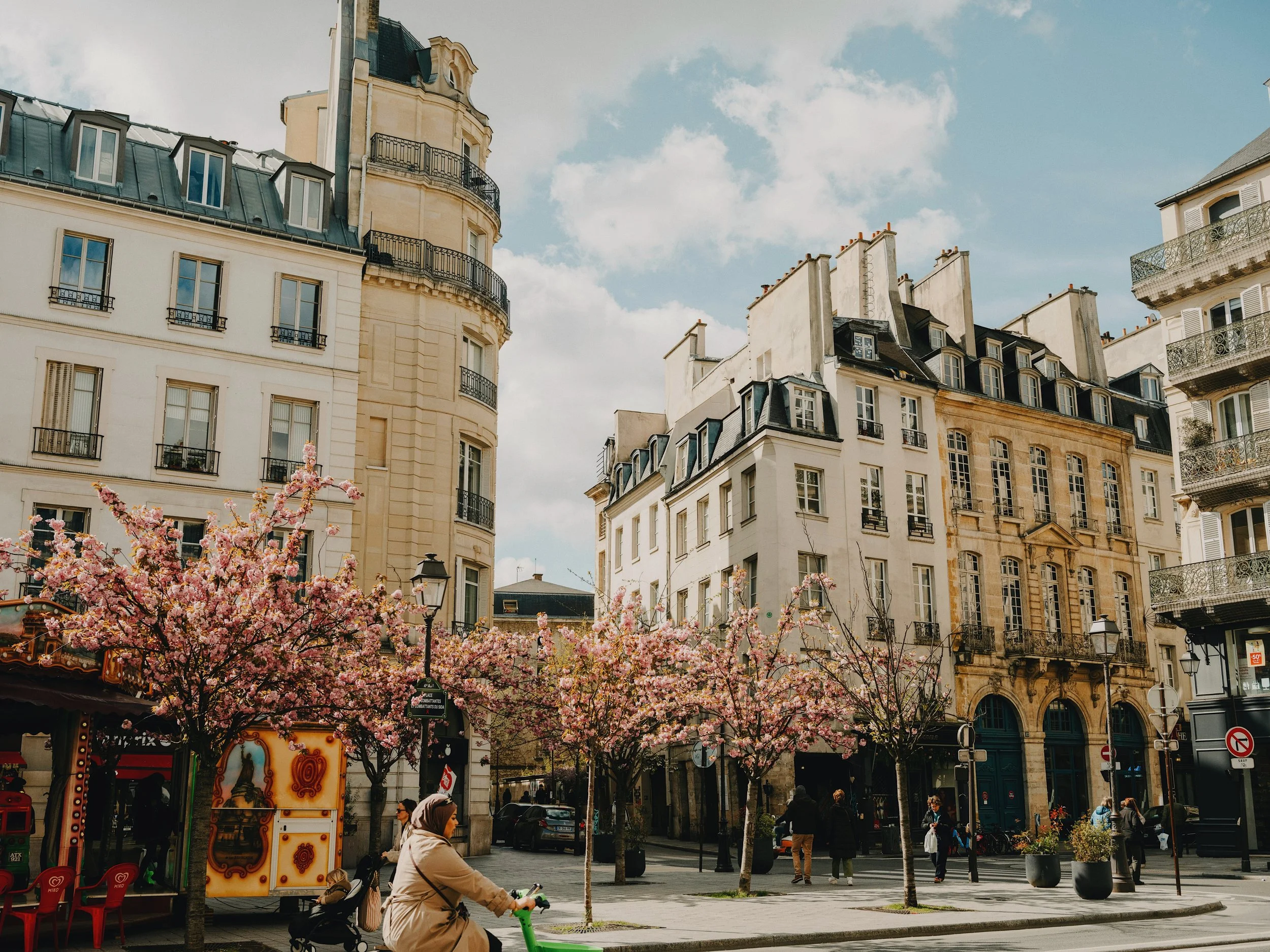 A Paris square in spring, cherry trees in bloom, Haussmann buildings behind, a cyclist passing in the foreground