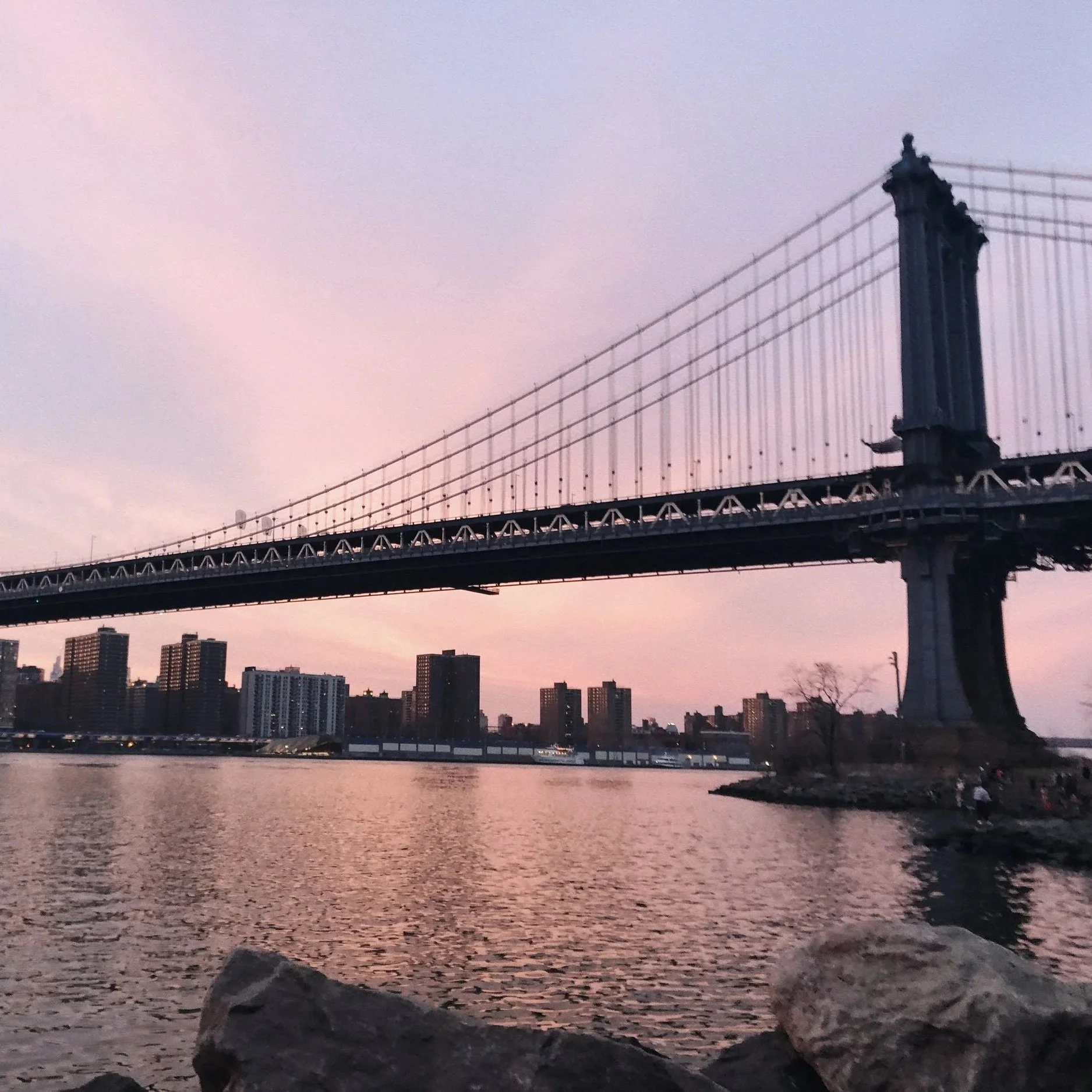 View of the Manhattan Bridge over the East River during sunset with an urban skyline in the background and rocks in the foreground.