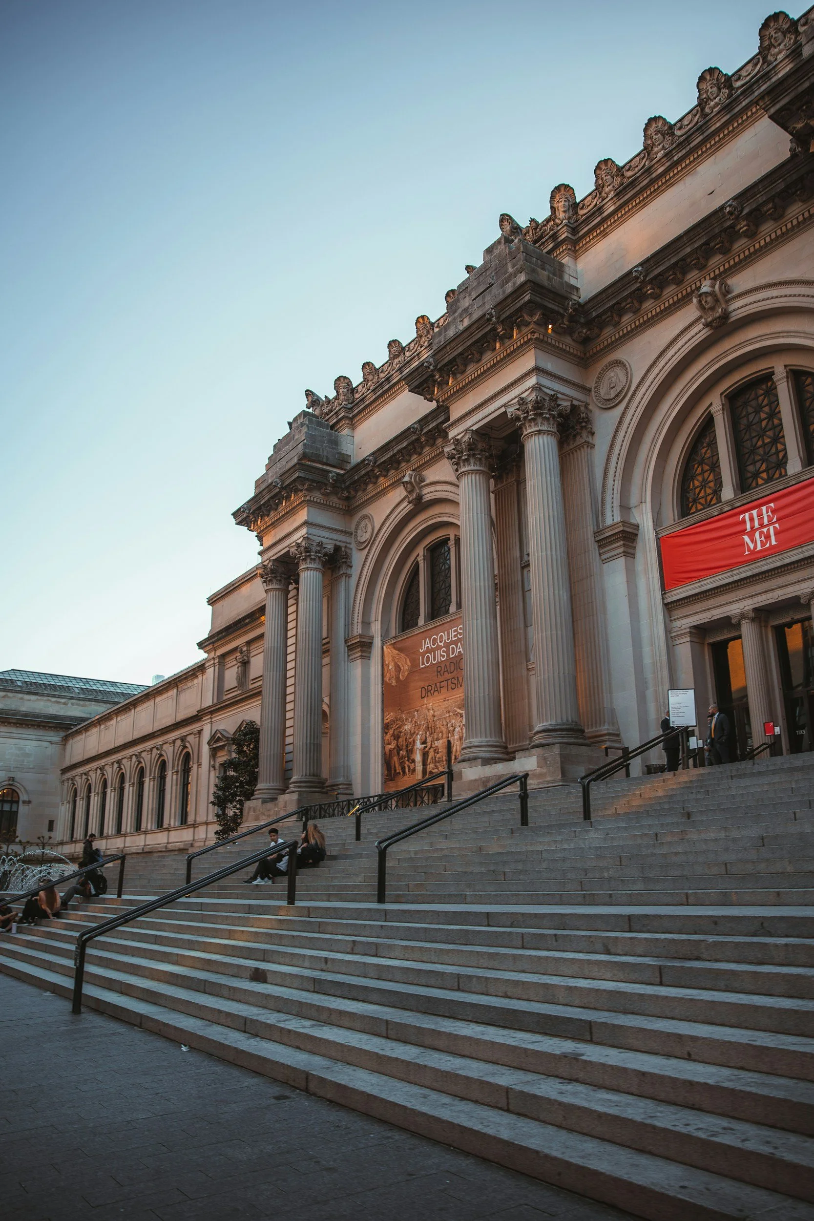 Exterior view of The Metropolitan Museum of Art, with stairs leading up to the entrance, columns, and banners for exhibitions, during early evening.