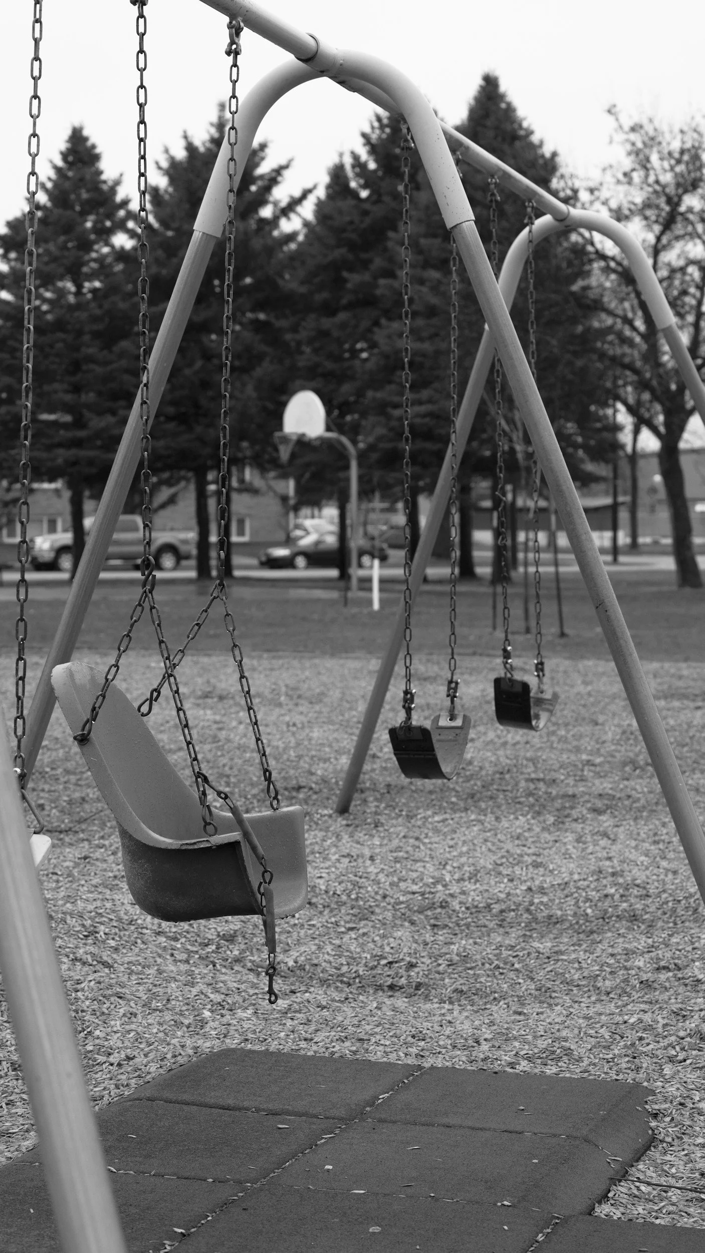 Empty swings at a playground with a gravel ground, trees, and a basketball hoop in the background. The image is in black and white.