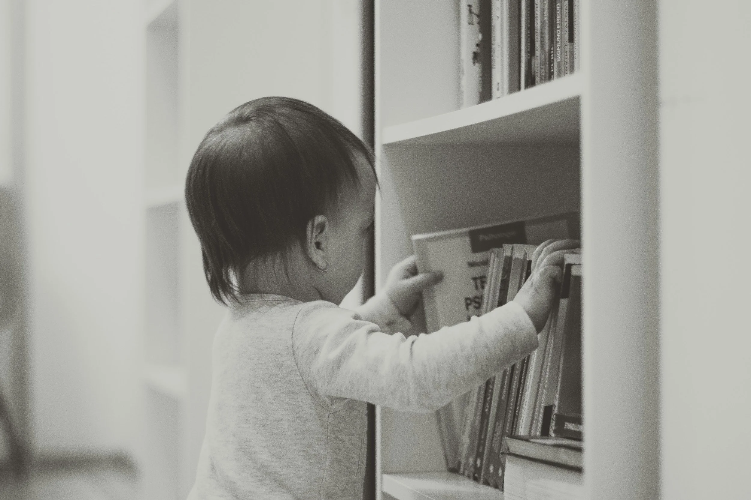 A young child with short hair reaching for a book on a bookshelf in a library or bookshop, in a black-and-white photo.