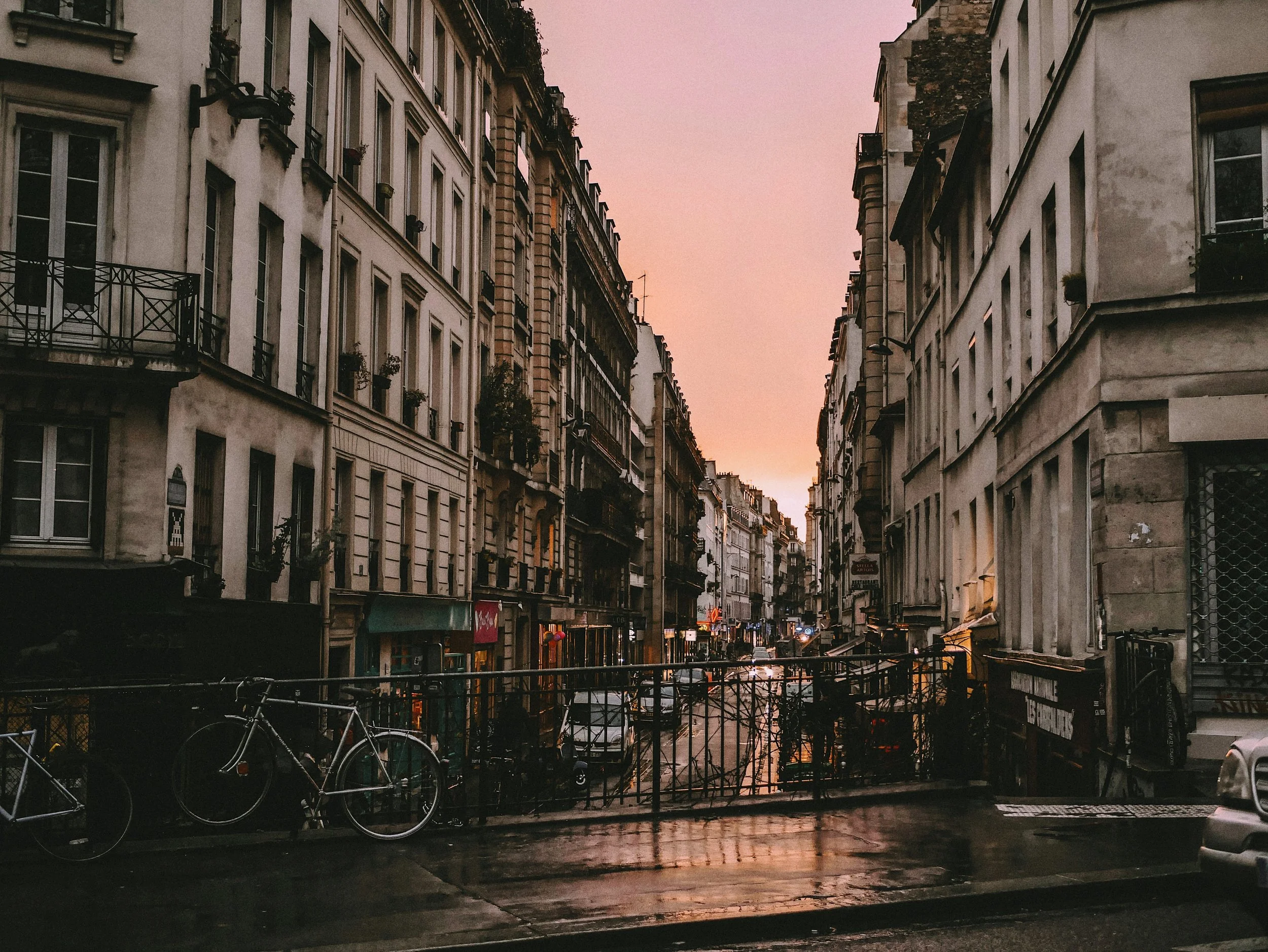 A rain-slicked Paris street at dusk, Haussmann facades rising on both sides, a bicycle locked to railings in the foreground