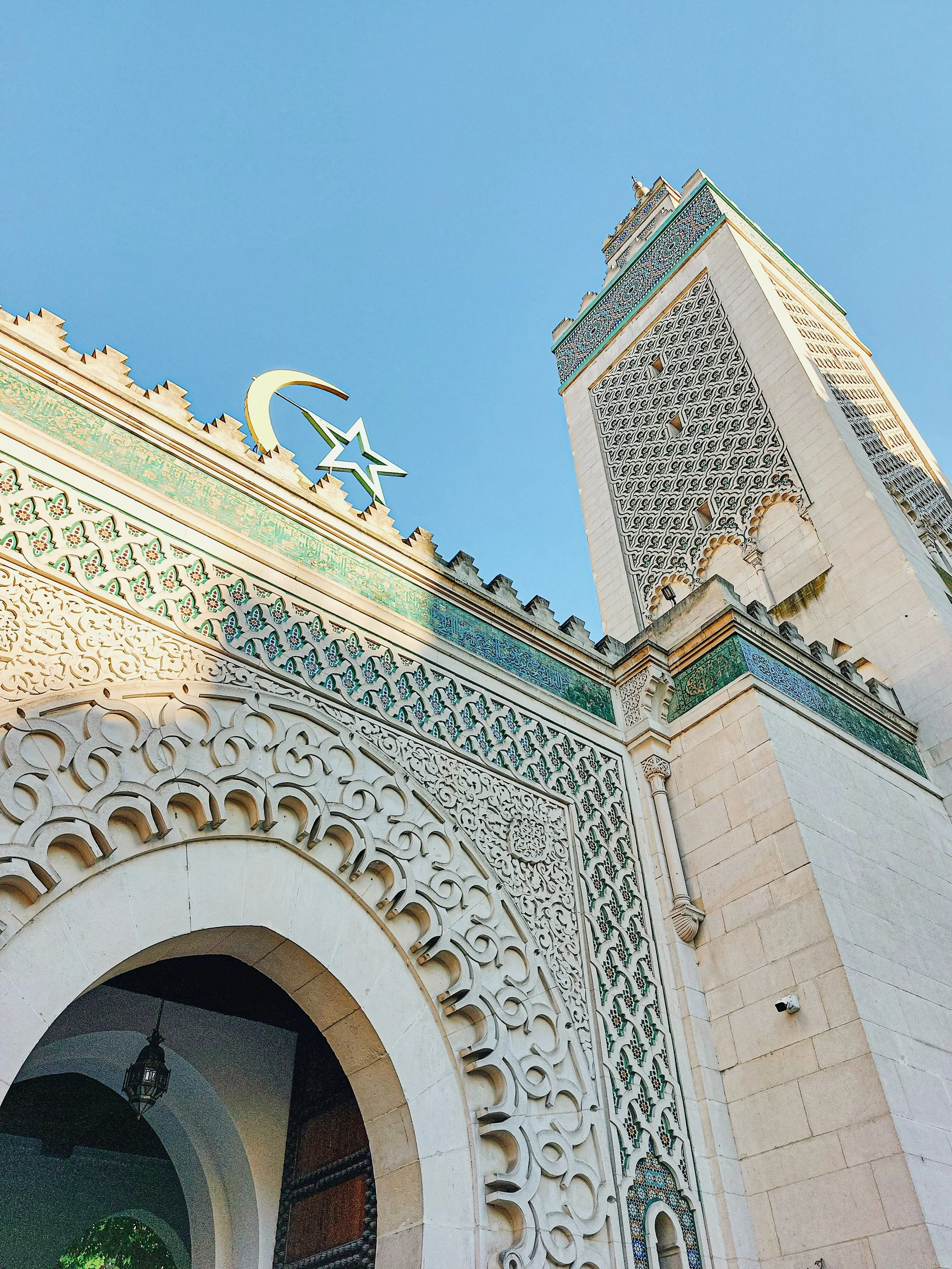 The ornate arched entrance and minaret of the Grande Mosquée de Paris, decorated with geometric tilework and a crescent and star, against a clear blue sky