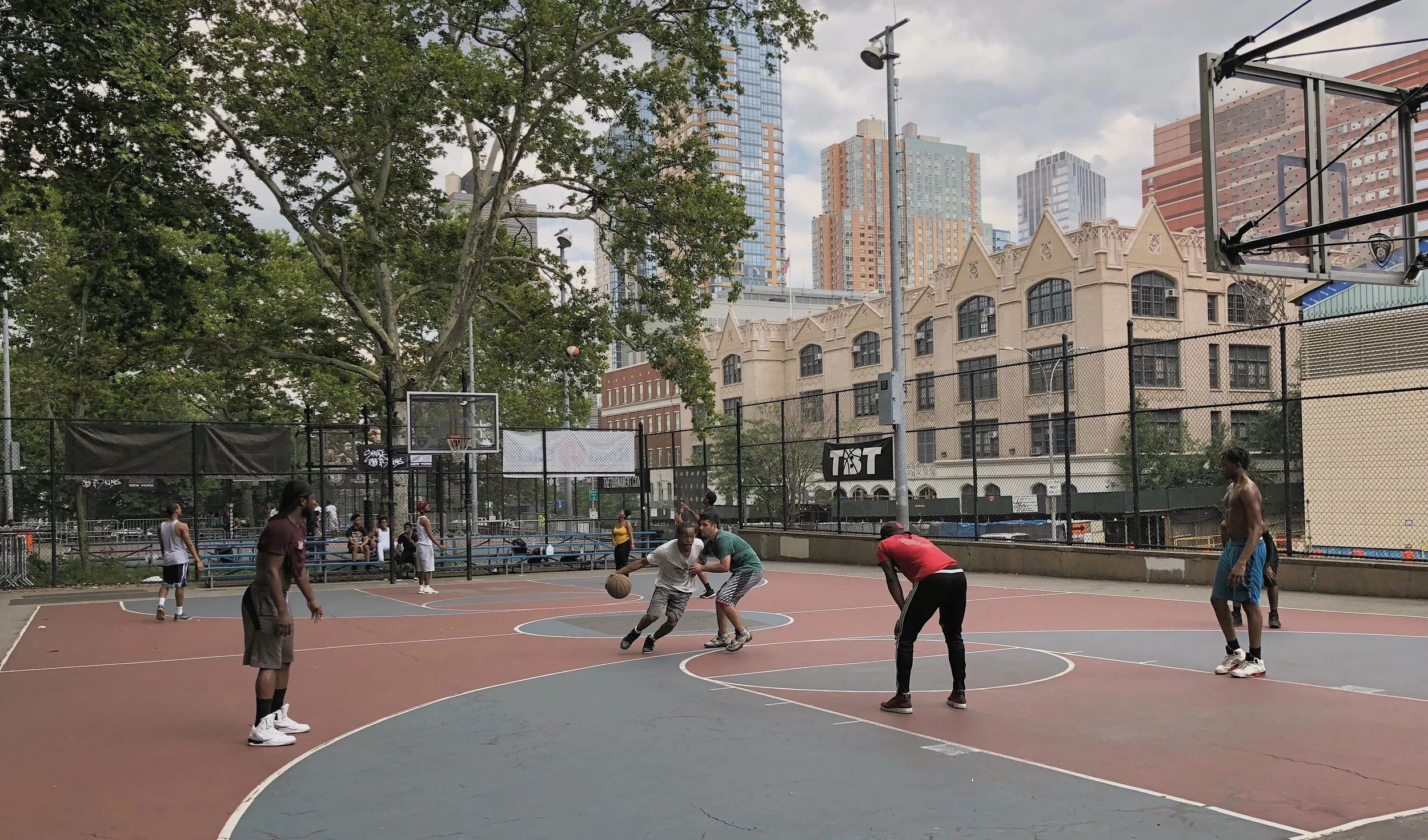 People playing basketball on an outdoor court with Manhattan skyline and historic buildings visible in the background
