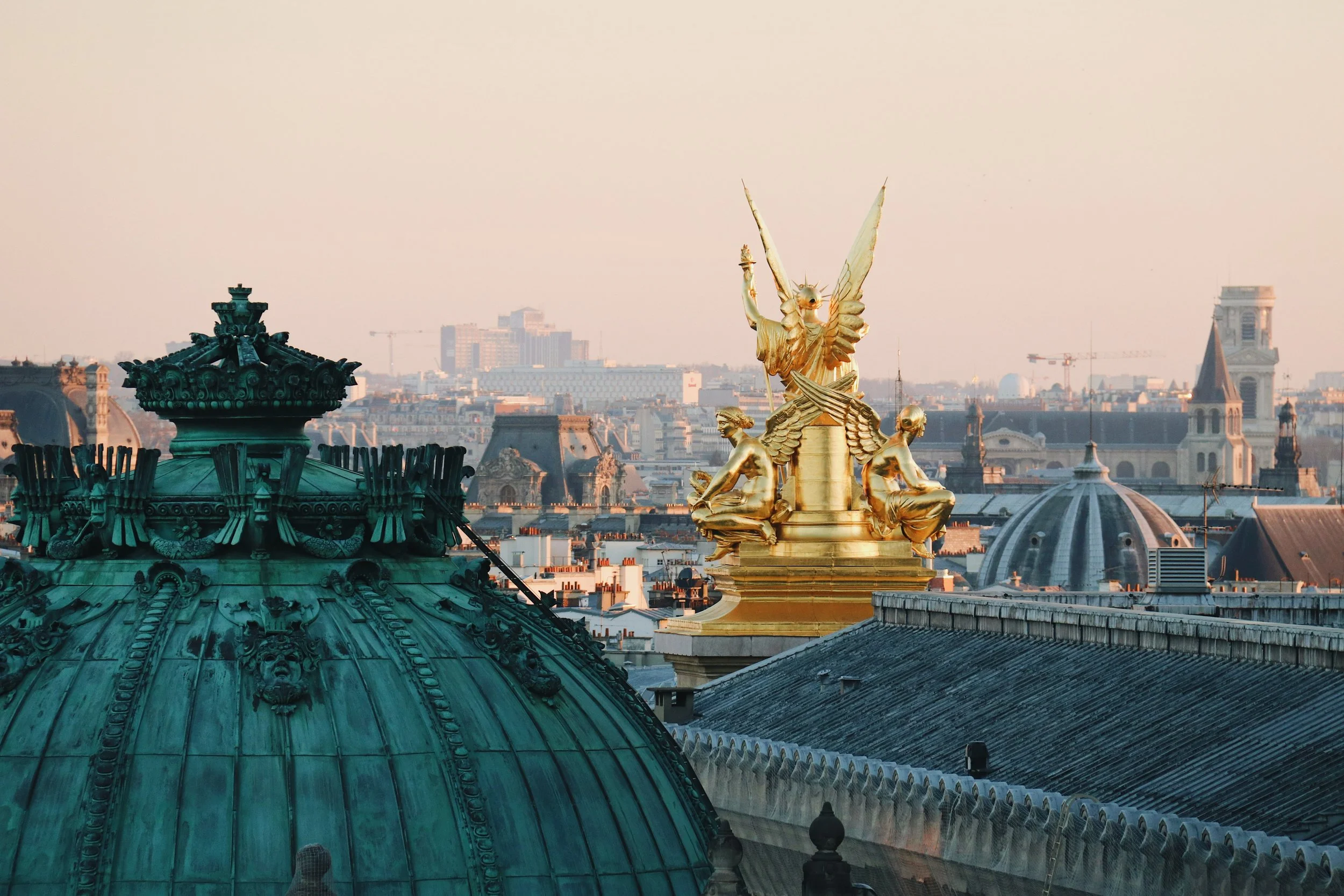 The gilded bronze sculpture atop the Opéra Garnier roof at dusk, the Paris skyline and Sacré-Coeur visible in the distance