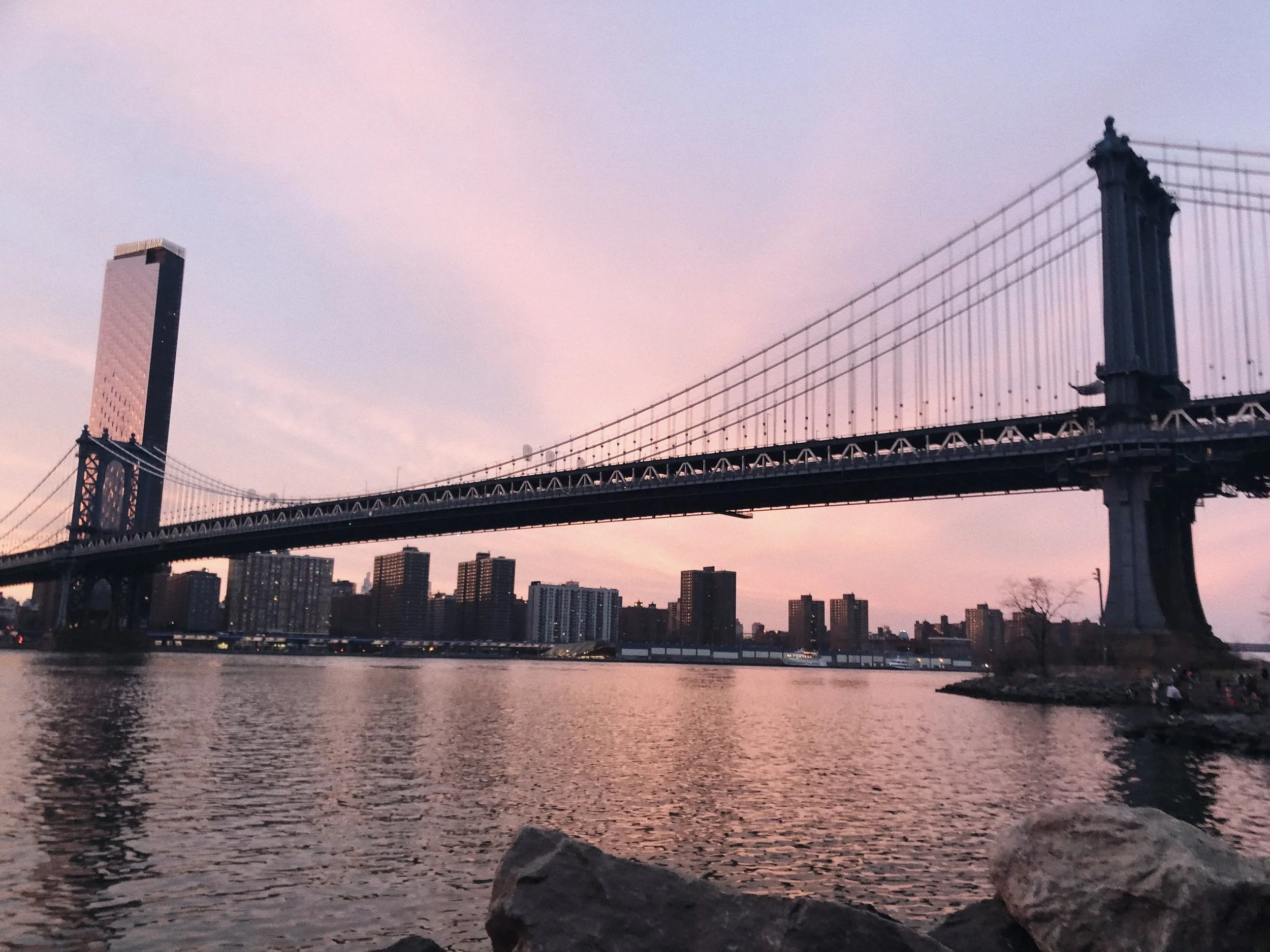 View of the Brooklyn Bridge spanning over the East River with the Manhattan skyline in the background during sunset. The sky is painted in pink and purple hues, and there are rocks in the foreground near the water.