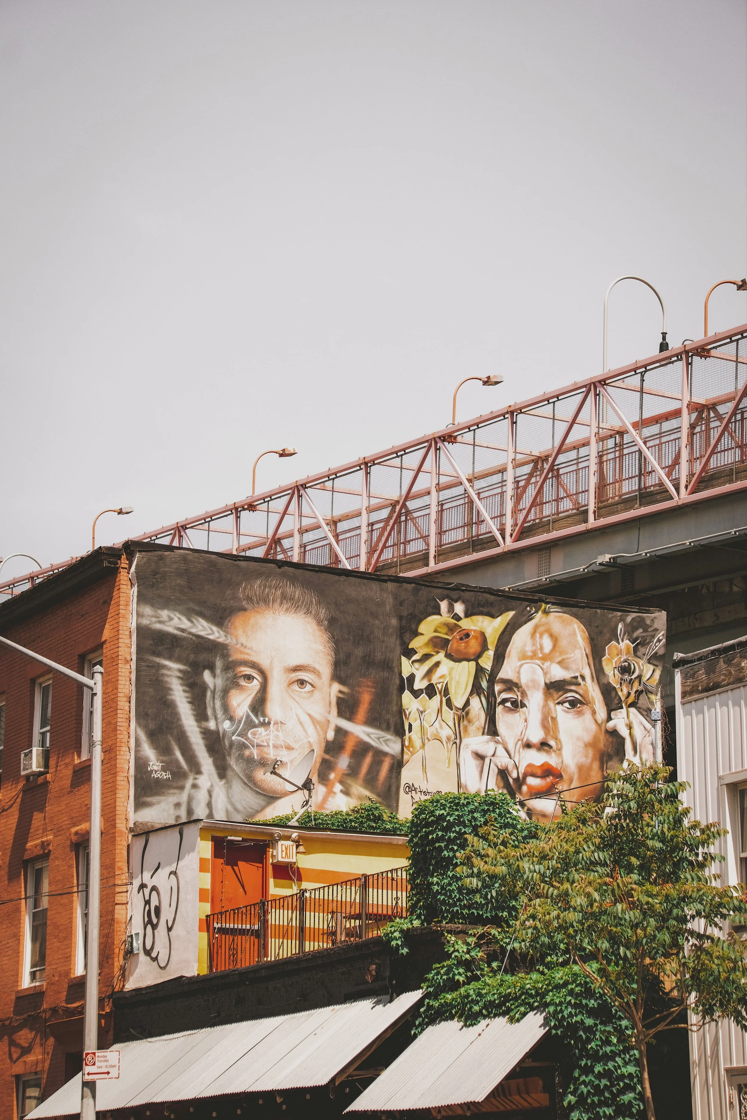 Large-scale street art mural depicting two faces on a building facade beneath an elevated bridge structure, Bushwick, Brooklyn