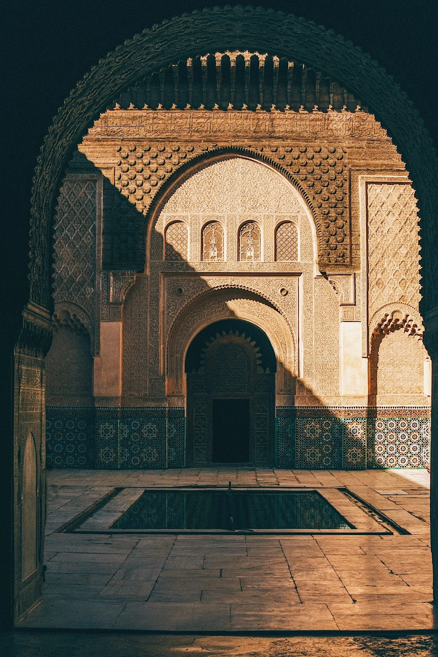 The carved stucco archway and zellij-tiled courtyard of Madrasa Ben Youssef in Marrakech, lit by warm afternoon light, a shallow reflecting pool at center