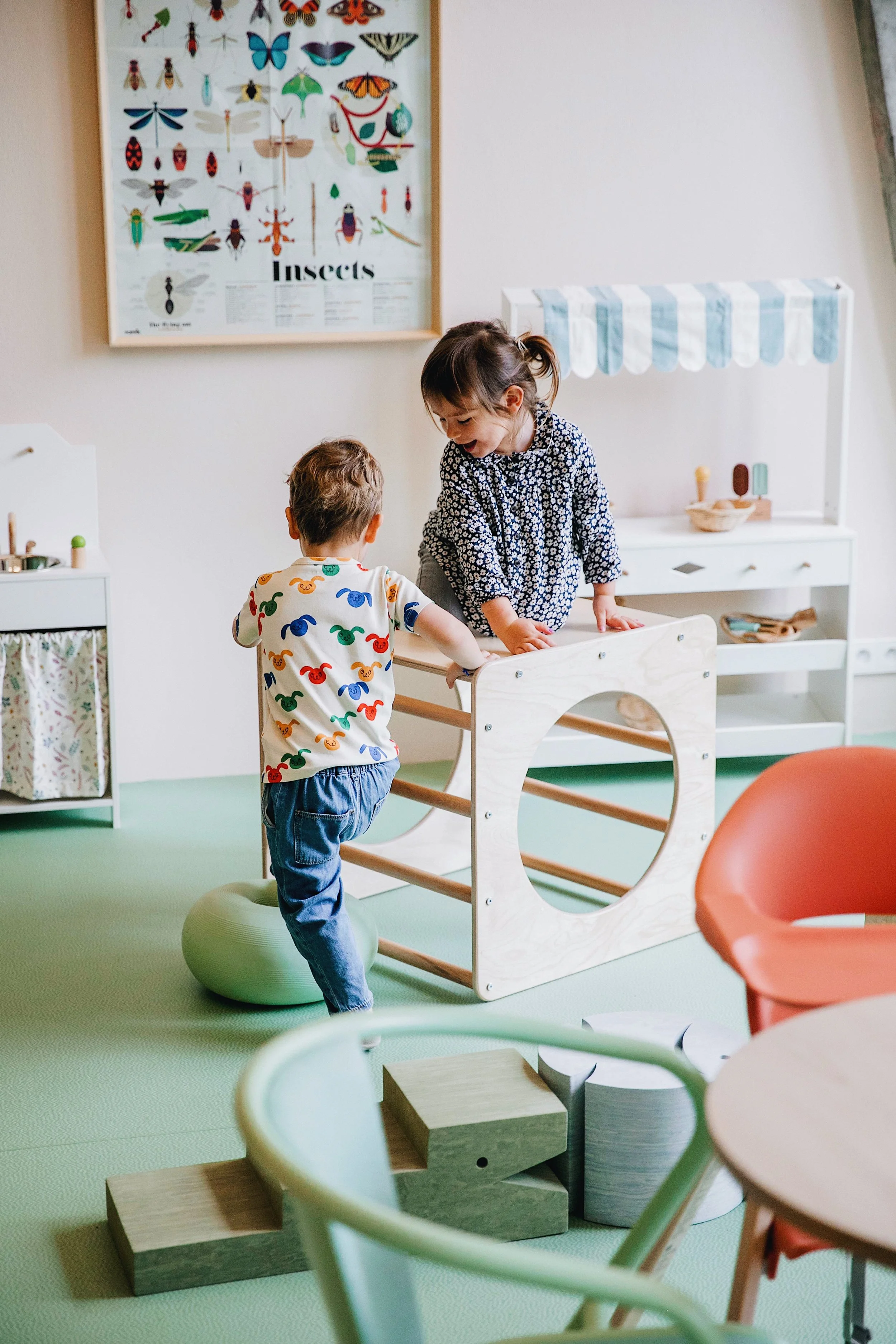 Two children playing on a wooden climbing frame at La Petite Forêt, a play café in Paris, with a play kitchen and insects poster in the background.