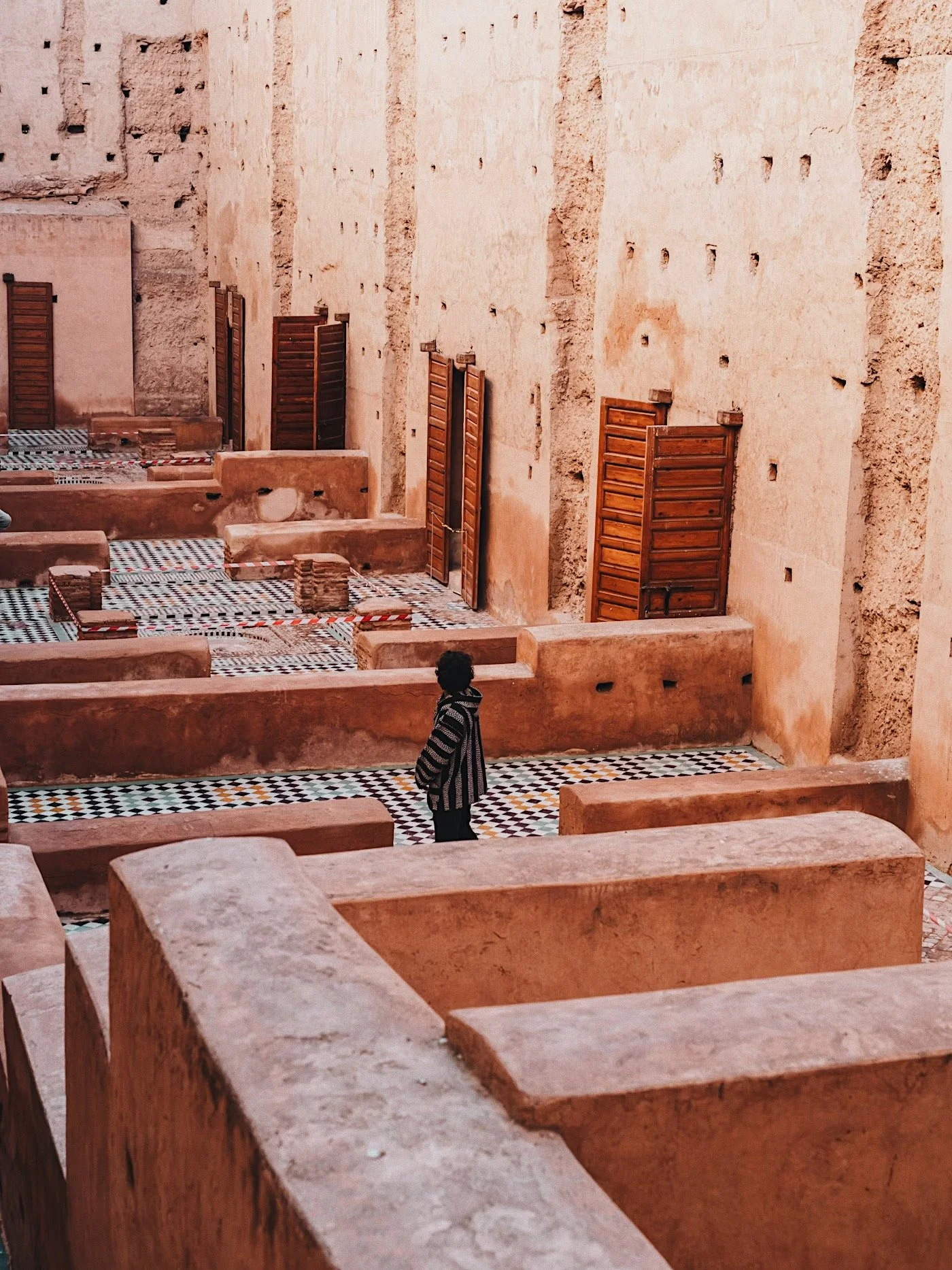 Stacked pink-earth medina rooftops in Marrakech, a figure in a striped djellaba standing on a terrace, wooden shutters and zellij tiling below
