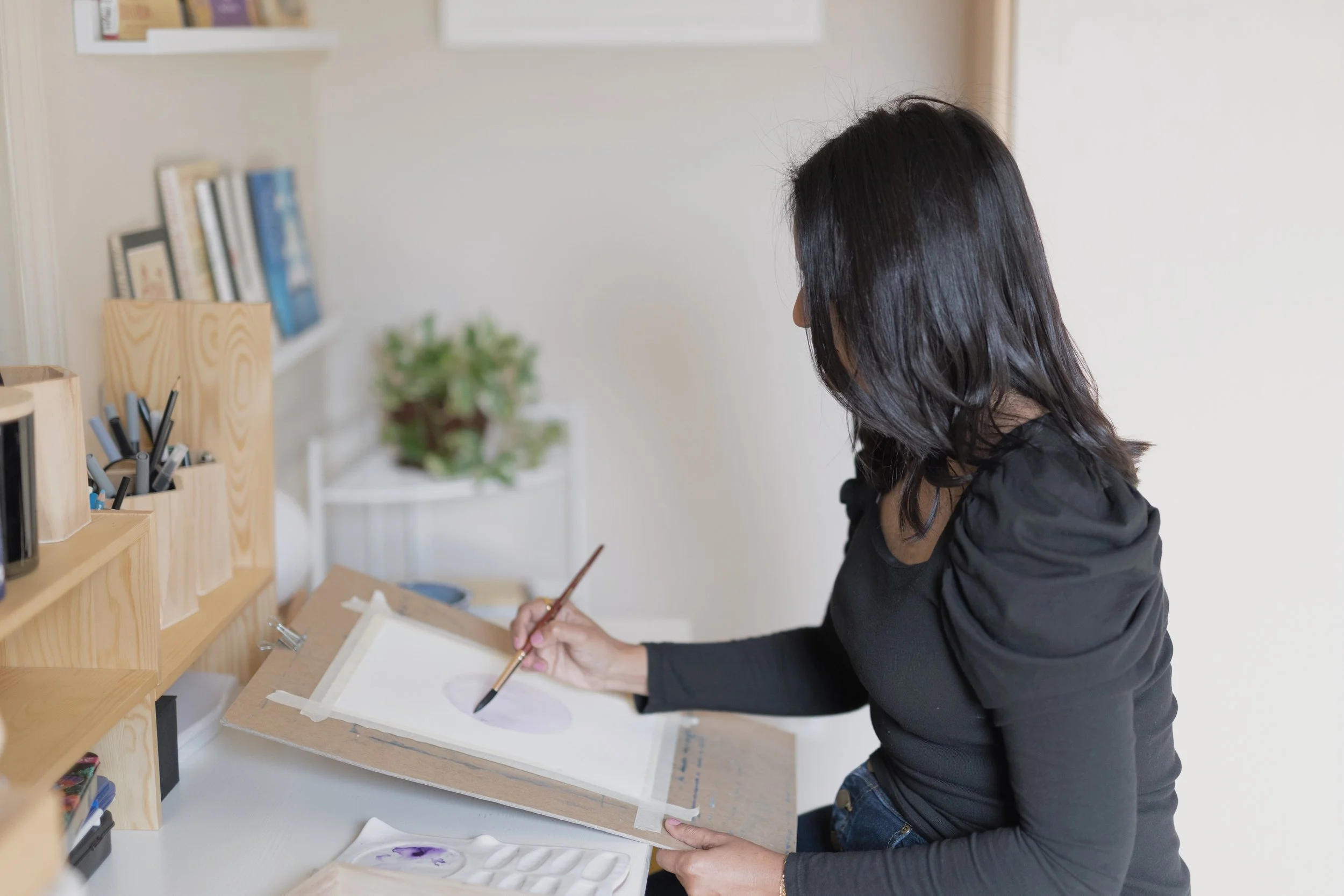 An illustrator seated at a desk painting on paper, viewed from the side.