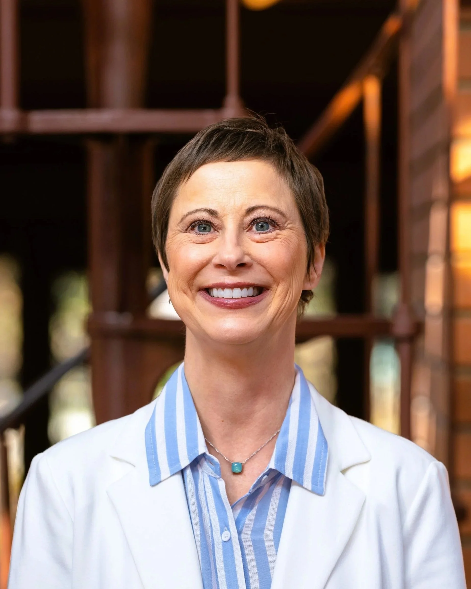 Woman sitting indoors smiling, wearing a black blazer over a white polka dot blouse, with a relaxed and comfortable atmosphere in the background.