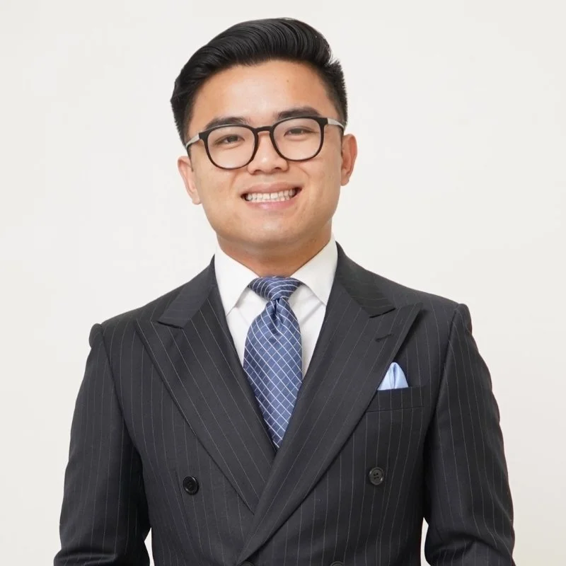 A young man wearing a dark pinstripe suit, white dress shirt, and a blue tie, smiling and looking at the camera.