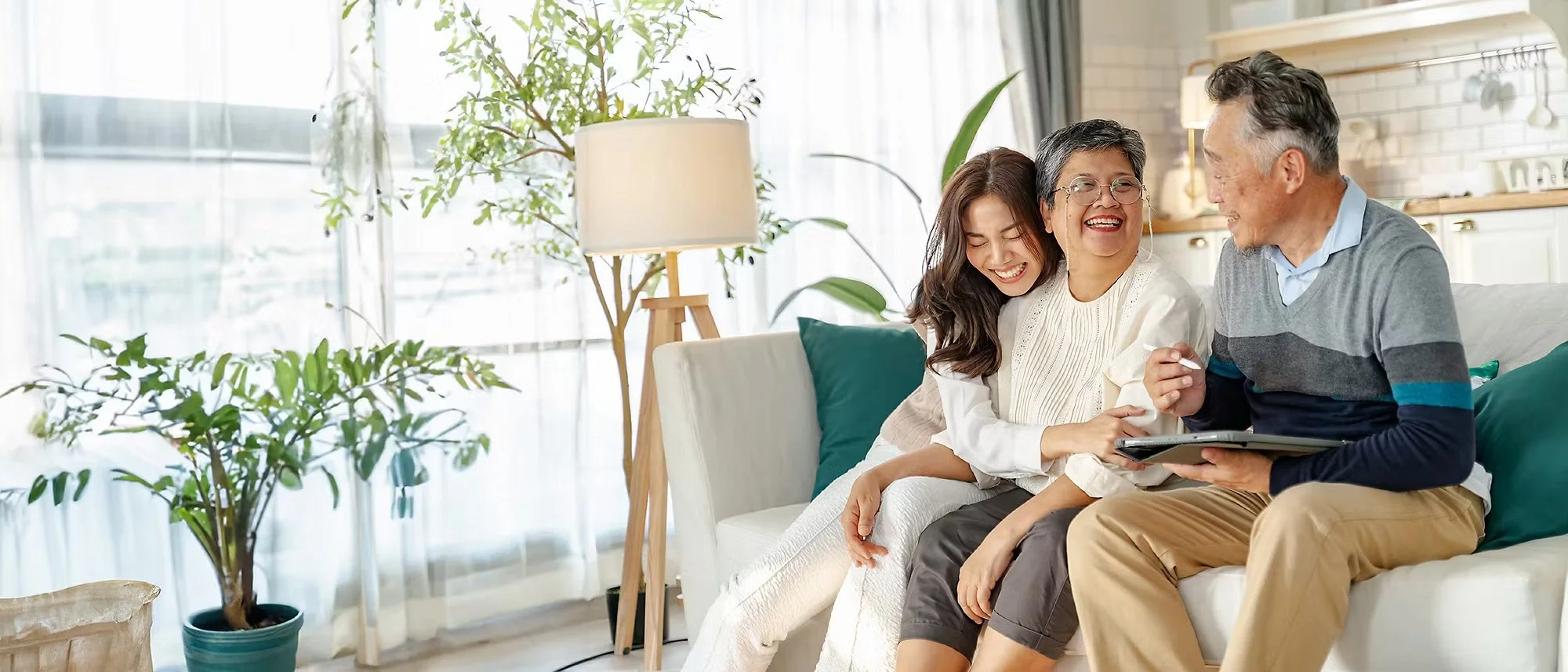 An elderly man and woman with a young woman sitting together on a sofa, smiling, looking at a tablet screen, in a bright living room with houseplants and natural light.