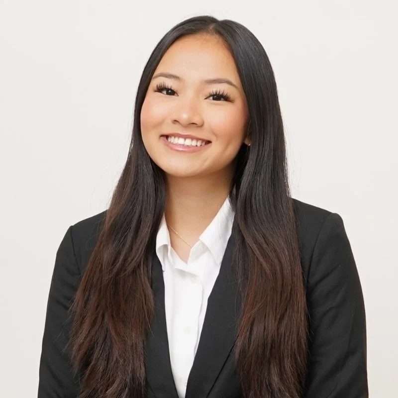 A young woman with long, dark brown hair, smiling, dressed in a black blazer and white shirt against a plain light-colored background.