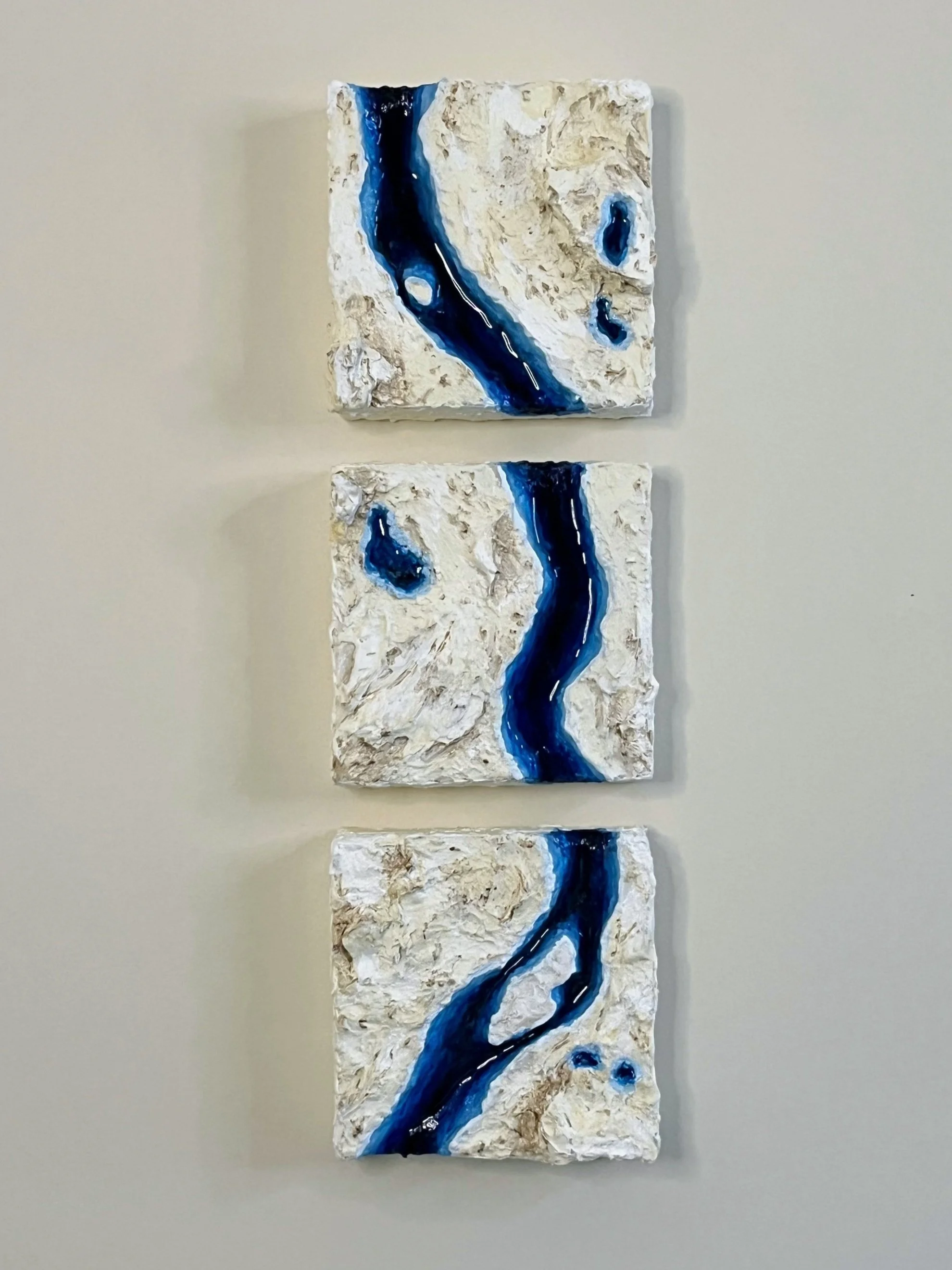 Wall art pieces featuring textured beige backgrounds with flowing blue river-like streaks.