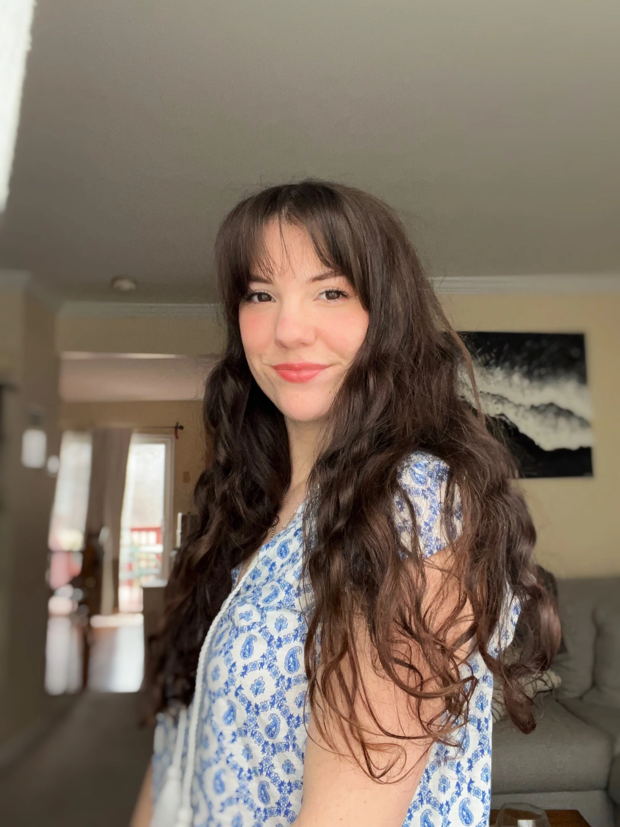 A young woman with long, wavy brown hair and a slight smile standing in a living room with natural light.