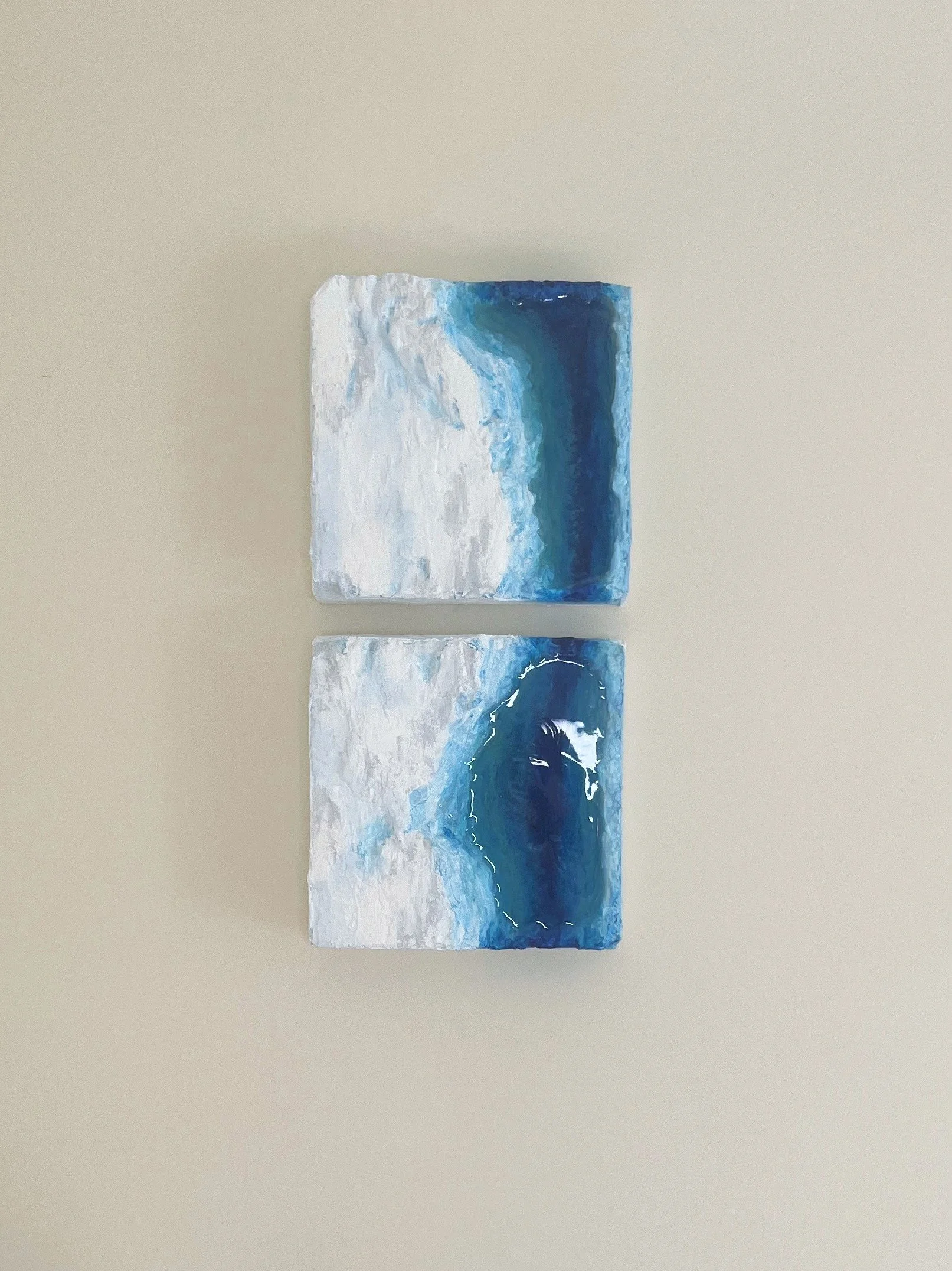 Two small square paintings of ocean cliff on a plain light-colored background, showing the water from different angles with textured white foam and blue water.