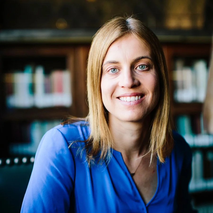 A woman with shoulder-length blonde hair and blue eyes smiling, wearing a blue blouse, in front of a bookshelf filled with books.