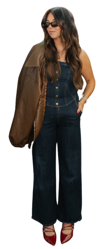 A woman with long wavy brown hair wearing sunglasses, a denim jumpsuit, a brown leather jacket draped over her shoulder, and red high heels.
